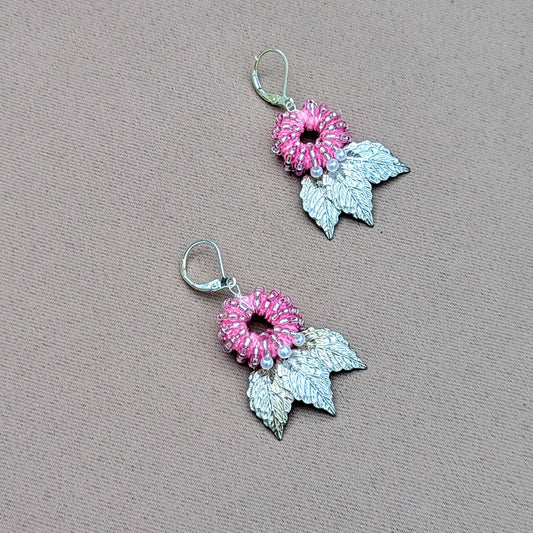 A unique handcrafted pair of earrings from the sustainable UK brand Mayaani Jewellery, featuring detailed silver leaves hanging beneath a circular pink beaded design with seed pearl accents. The earrings showcase intricate craftsmanship with a natural, eco-friendly aesthetic.