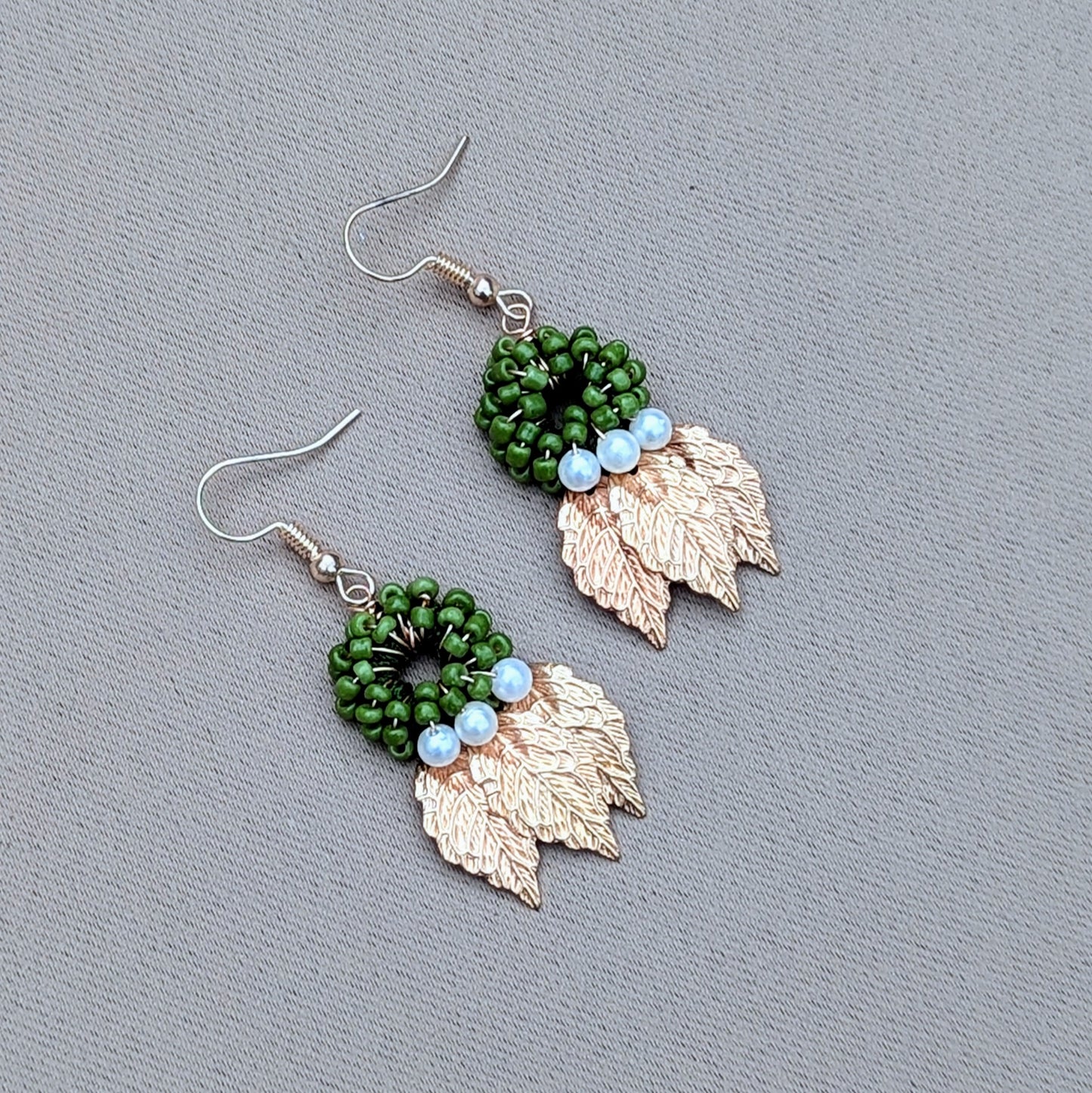A uniquely artisan handcrafted pair of earrings from sustainable UK brand Mayaani Jewellery, featuring a circular green beaded design with gold leaves dangling beneath and seed pearl accents. This pair showcases intricate craftsmanship with a natural nature loving aesthetic.