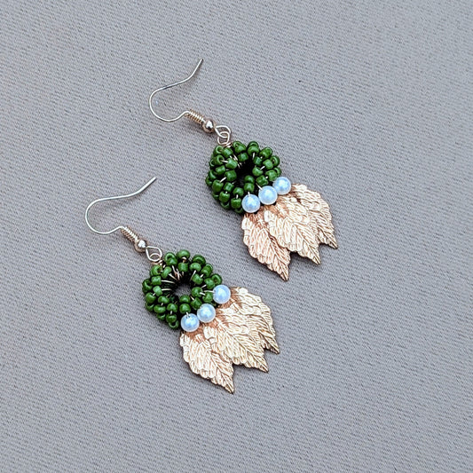 A uniquely artisan handcrafted pair of earrings from sustainable UK brand Mayaani Jewellery, featuring a circular green beaded design with gold leaves dangling beneath and seed pearl accents. This pair showcases intricate craftsmanship with a natural nature loving aesthetic.