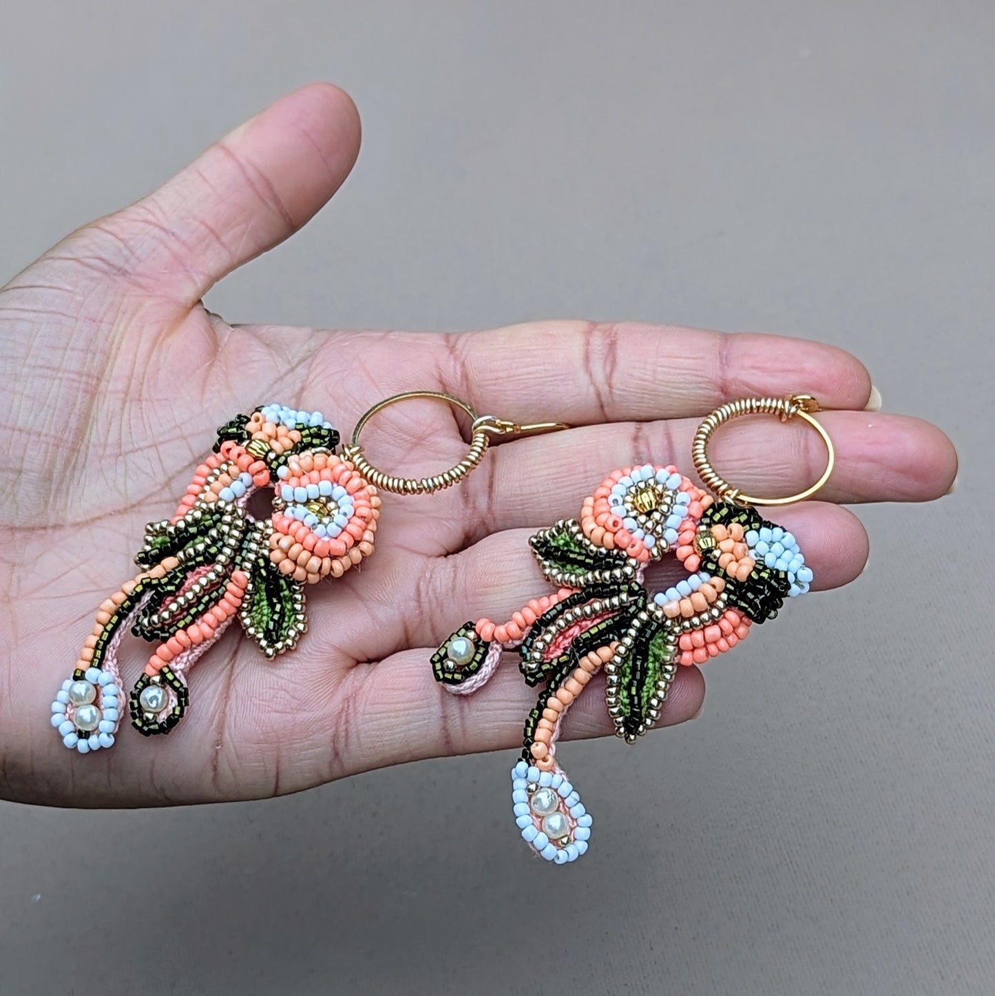 Enhance your collection with these sustainably handcrafted Indian statement earrings pictured in the palm of a hand. Adorned with green gold, peach, and white beads, they showcase hand wire-wrapped gold hoops and refined seed pearl details. Exclusively available from Mayaani Jewellery, a UK brand that specializes in comfortably lightweight, sustainable jewellery for Indian weddings and special events.