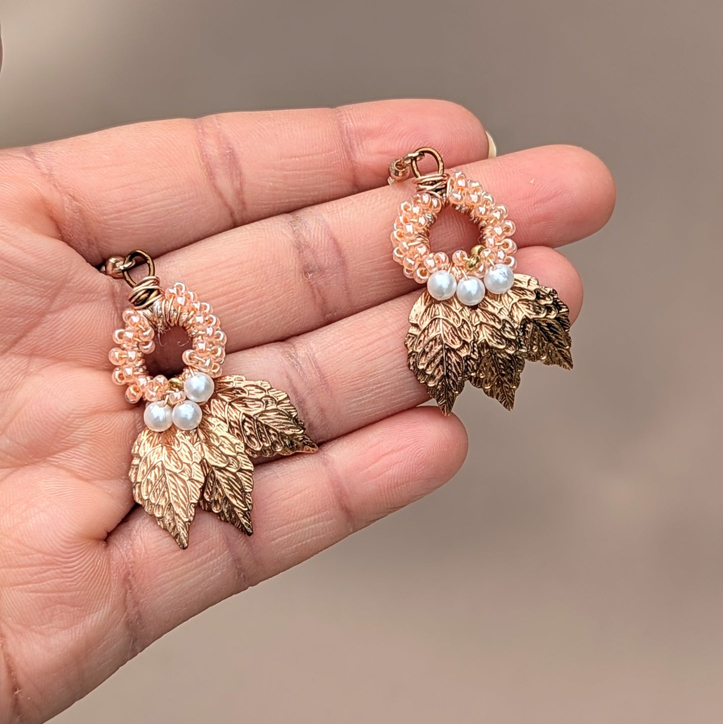 Hand holding a handcrafted pair of Indian Boho-style earrings from sustainable UK brand Mayaani Jewellery. Featuring gold-plated leaves and intricate hand crochet. The earrings combine a loop of peach beads, delicate white seed pearls, and detailed hand wire bead work. This limited edition best selling design embodies a fusion of traditional artistry and modern fashion made with sustainable materials by a UK-based brand.