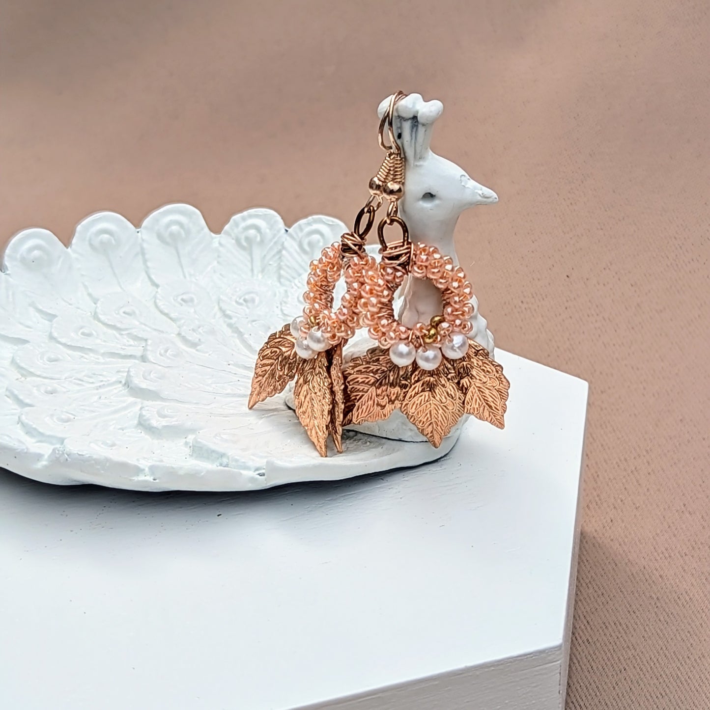 A pair of artisan handcrafted earrings by sustainable brand Mayaani Jewellery UK displayed on a white peacock trinket dish. Featuring gold-plated leaves, hand-crocheted elements and white seed pearl accents. These limited edition earrings incorporate detailed wire work and beadwork. The perfect touch for everything from occasion to everyday wear.