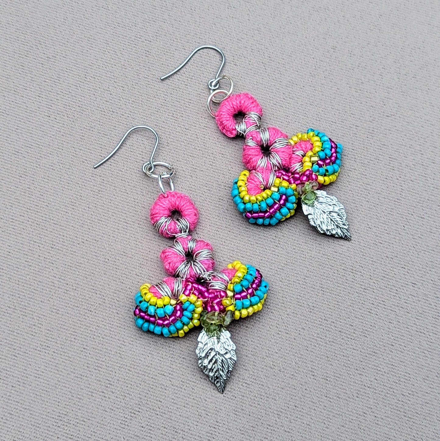 A close-up of a one-of-a-kind of handcrafted earrings by sustainable UK brand, Mayaani Jewellery. These Indian-inspired earrings feature vibrant pink, yellow, turquoise, and purple crochet work, adorned with intricate beading and finished with silver leaf charms. They are exceptionally lightweight and made from high-quality upcycled materials, including peridot gemstone chips, reflecting the brand's dedication to sustainability, artistry and comfort.