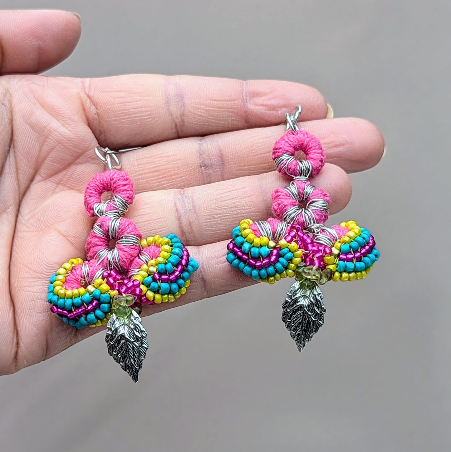 Hand holding an Indian-inspired pair of earrings designed by sustainable UK brand Mayaani Jewellery. The earrings feature a colourful combination of pink crochet, multi-hued beads, and silver leaf charms. Crafted with upcycled materials, including peridot chips, they are incredibly lightweight and comfortable, highlighting Mayaani's dedication to sustainability and artisanal craftsmanship with a vibrant, eco-conscious design.