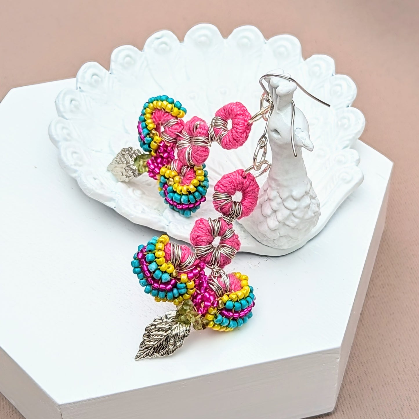 Displayed on a white peacock trinket dish and handcrafted by the sustainable UK brand Mayaani Jewellery, these Indian-inspired earrings feature a vibrant mix of colours with intricate pink crochet, multi-coloured beadwork, and silver leaf charms. Made from upcycled materials, including peridot chips, they are lightweight and eco-conscious. Their bold yet delicate design embodies Mayaani's focus on sustainability, artistry and high-quality craftsmanship.