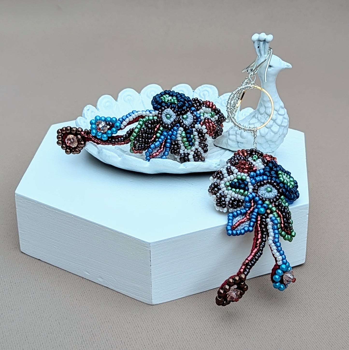 A bold vibrant pair of Indian-inspired earrings featuring intricate floral inspired beadwork and gold and silver accents displayed on a white peacock trinket dish. Exclusively from Mayaani Jewellery UK, this beautiful statement design showcases artistic craftsmanship steeped in Indian culture with a modern edge.