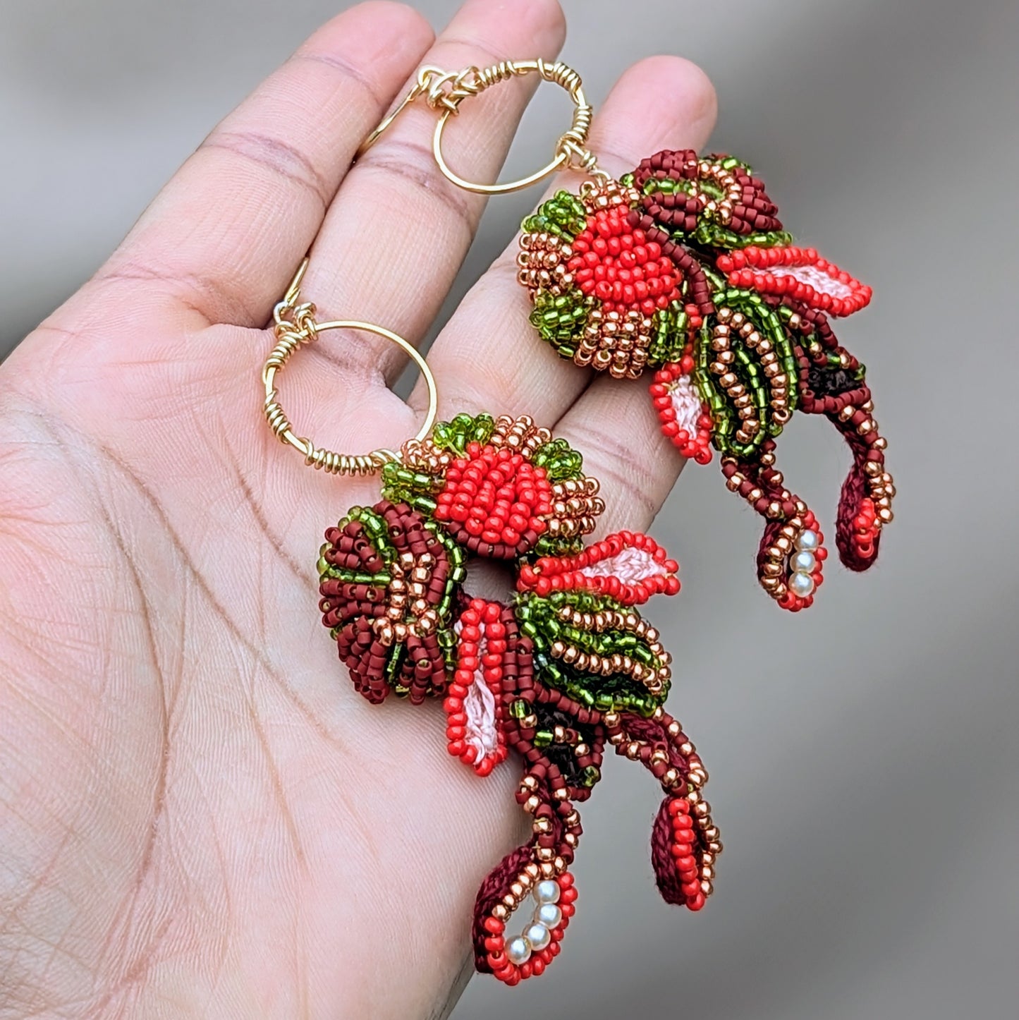 A hand holding a pair of handcrafted Indian inspired earrings exclusively from Mayaani Jewellery UK, a sustainable online wedding and occasion jewellery brand. Featuring exceptional bead artistry in red, green, gold and brown, they are stylish and comfortable hanging on a hand wire wrapped gold hoop. They embody the brand's focus on sustainability and craftsmanship.