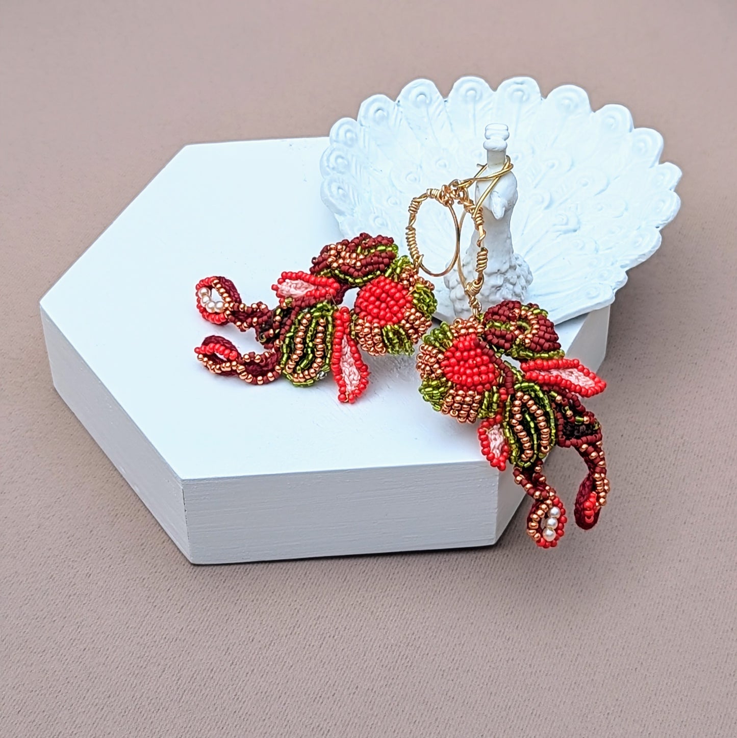 A peacock trinket dish showcasing a pair of handcrafted statement beaded earrings designed and handcrafted exclusively for Mayaani Jewellery. Featuring artistic beadwork in red, brown, green and gold on a wire wrapped gold hoop. Inspired by Indian culture and heritage they offer a perfect blend of cultural elegance and comfort.