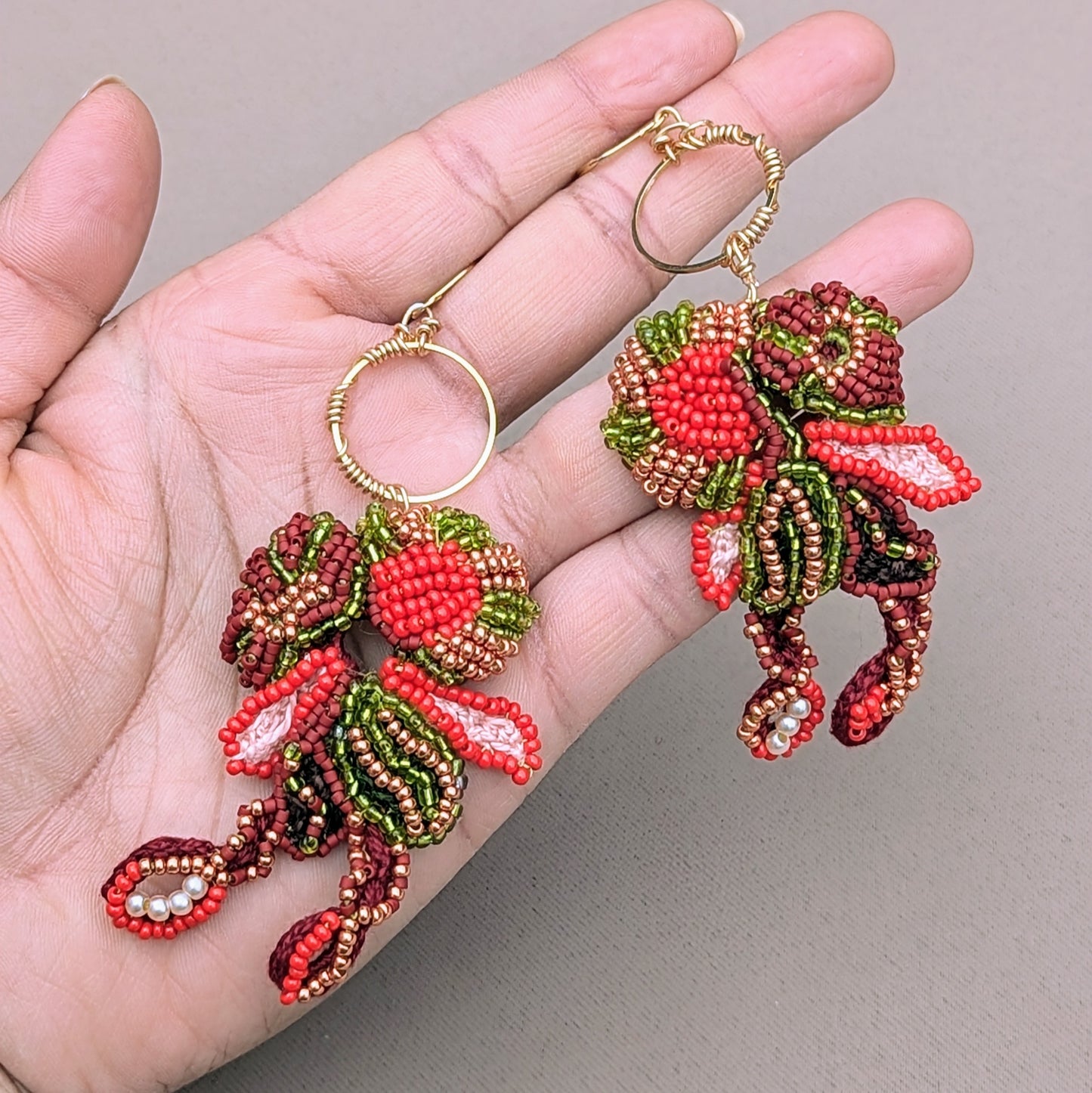 A hand holding a beautiful pair of earrings, crafted for UK sustainable brand Mayaani Jewellery, showcasing detailed red, green and gold floral and leaf motif beadwork. The bold design speaks to the richness of Indian culture and heritage whilst being comfortable to wear.