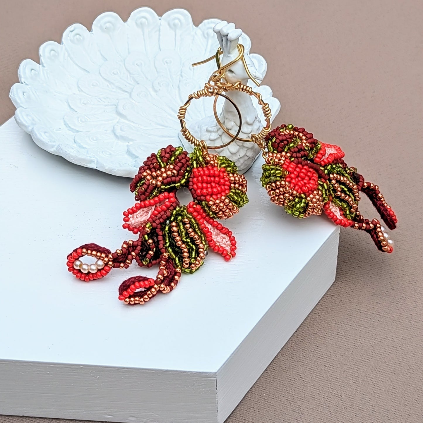 Displayed on a white peacock trinket dish, a stunning pair of Indian-inspired earrings, made for Mayaani jewellery, featuring detailed beadwork in red, green, gold and brown dangling on a gold wire wrapped hoop. Crafted by UK designer Megha, the earrings are versatile, comfortable, and reflect the brand's dedication to sustainability and expert craftsmanship.