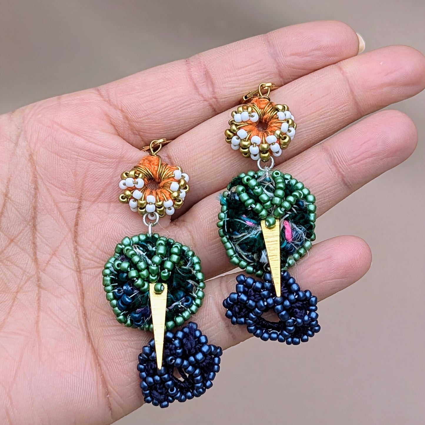 Hand holding a pair of statement beaded Indian style artisan earrings against a neutral backdrop. Exclusive to Mayaani Jewellery this pair features the brand's signature scrap beads and 18K gold triangle charms in a blend of white, green, gold, blue and orange bead, crochet and wire work.