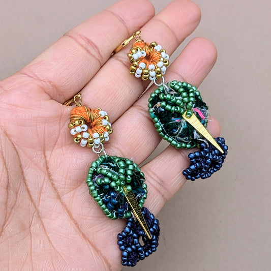 Hand holding a tree of life Akshaya Vata pair of blue, green, orange, gold and white bead crochet earrings from sustainable UK brand Mayaani Jewellery. Featuring gold plated stainless steel ear clasps, 18K gold triangle charms and signature waste recycling scrap beads.