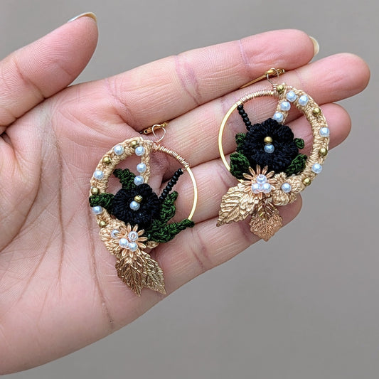 Artisan-crafted Aloki earrings displayed in hand, featuring black and gold floral crochet with brass and seed pearl details.