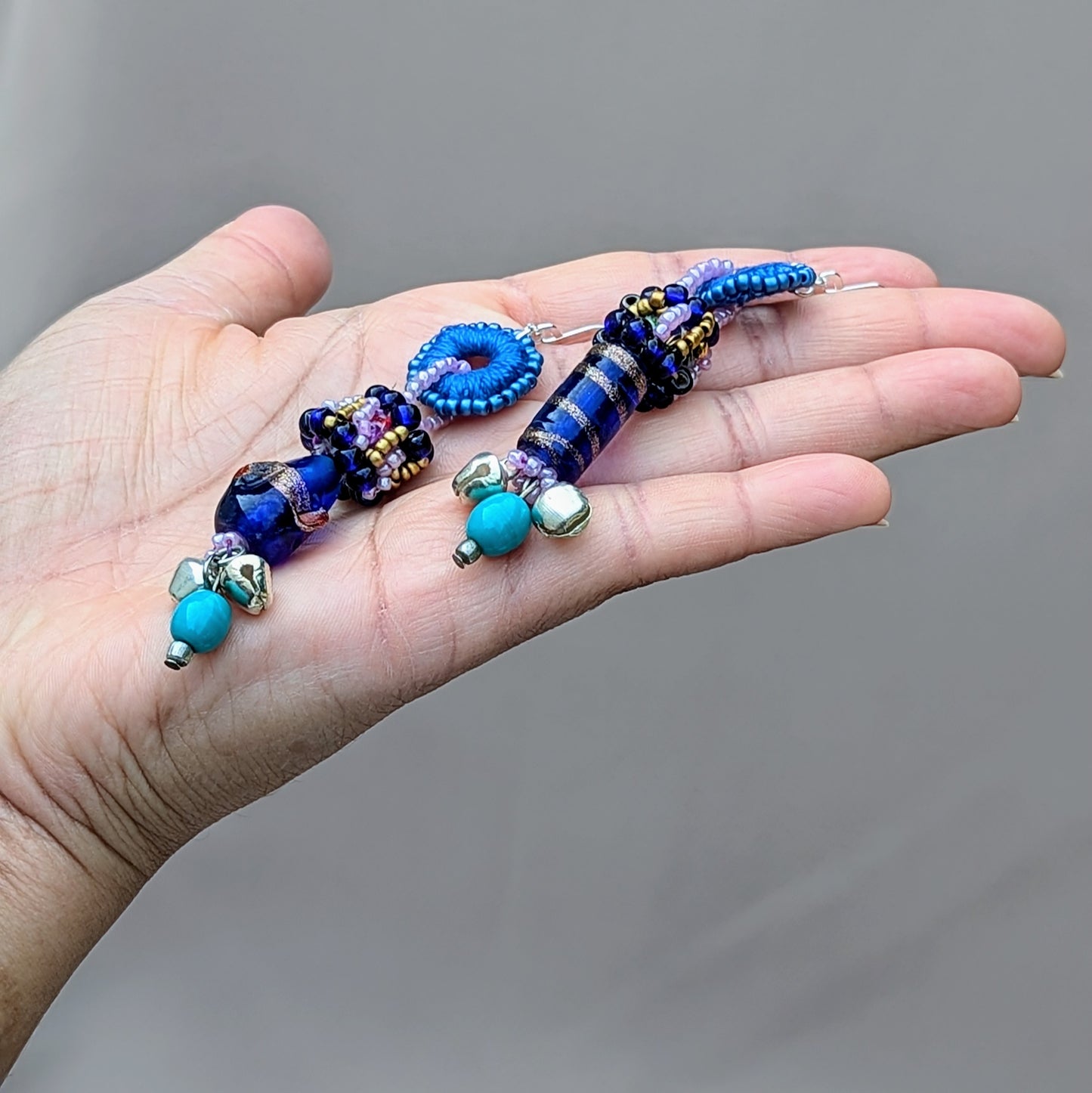 Asymmetric blue and silver statement earrings resting on the palm of a hand, fine handcrafted details visible.