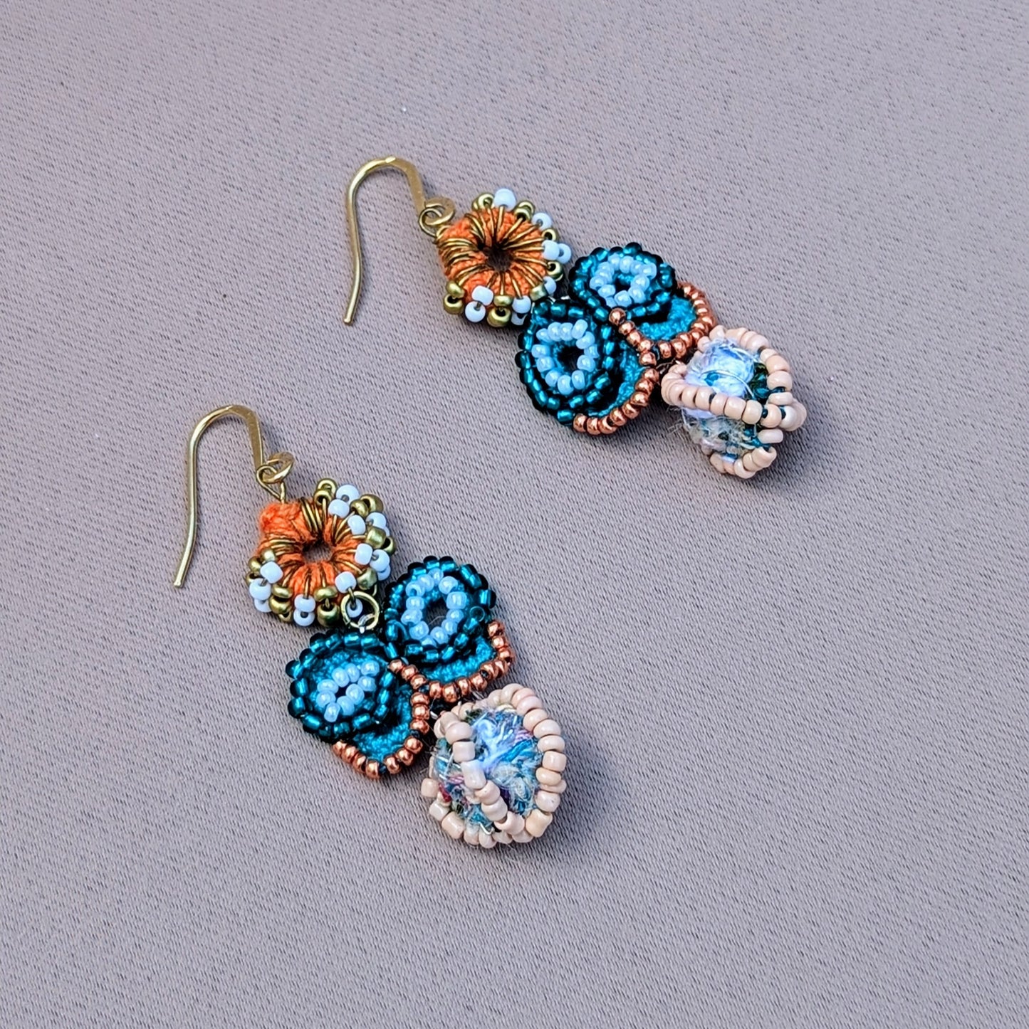 Eco-friendly butterfly earrings handcrafted by Mayaani Jewellery UK, featuring a turquoise blue and rose gold seed bead butterfly, orange crochet circle with white and gold accents, and a unique zero-waste scrap bead drop. Finished with gold heavy duty stainless steel hooks.