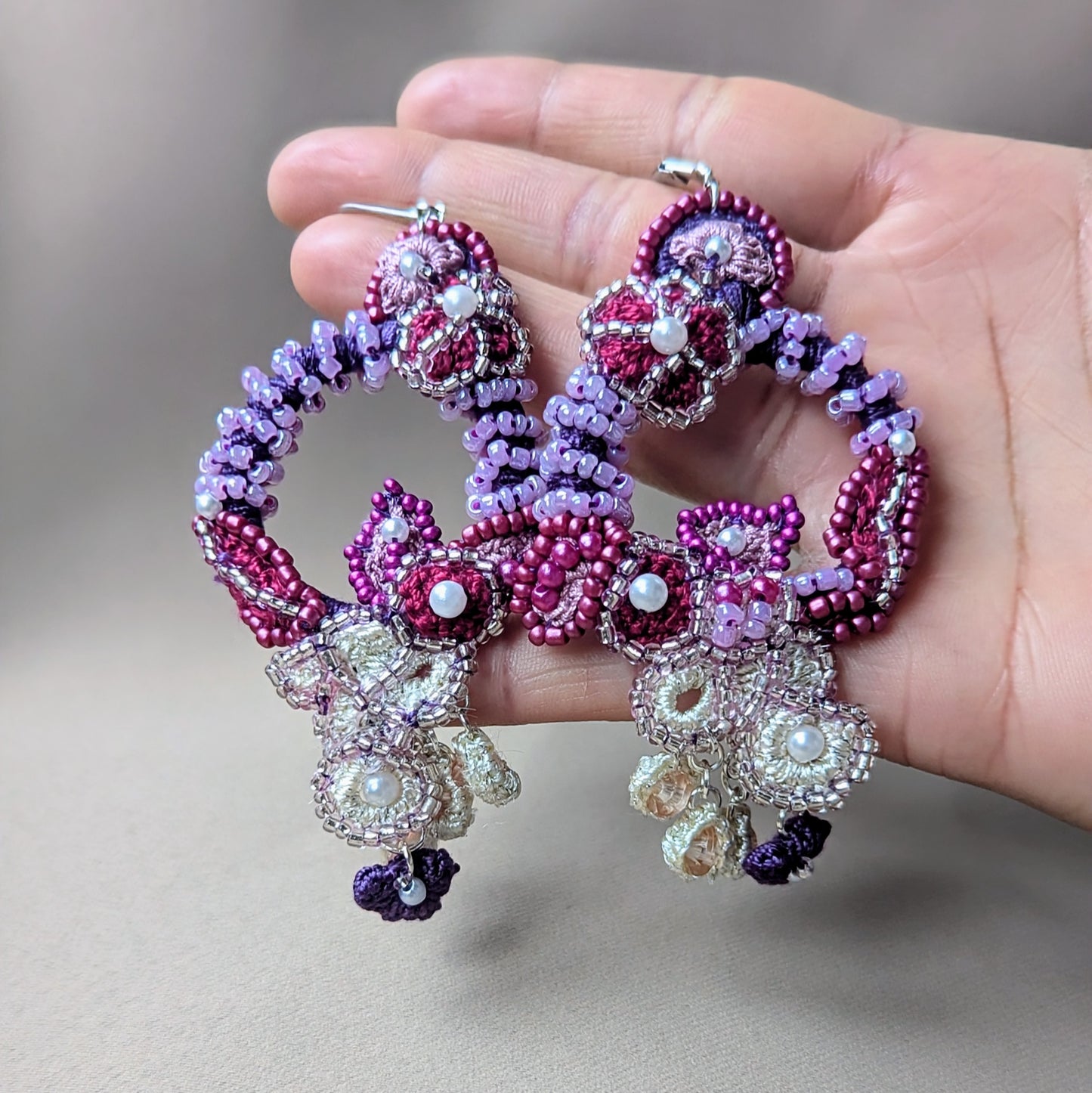Hand holding the statement earrings from the 'Gulab Rani' Indian bridal jewellery set by Mayaani Jewellery UK. These intricately handcrafted earrings feature sculptural bead embroidery and crochet work in shades of rich red, lavender, ivory and deep plum, with floral motifs and cascading bead details. A bold, elegant piece designed to celebrate individuality and heritage.