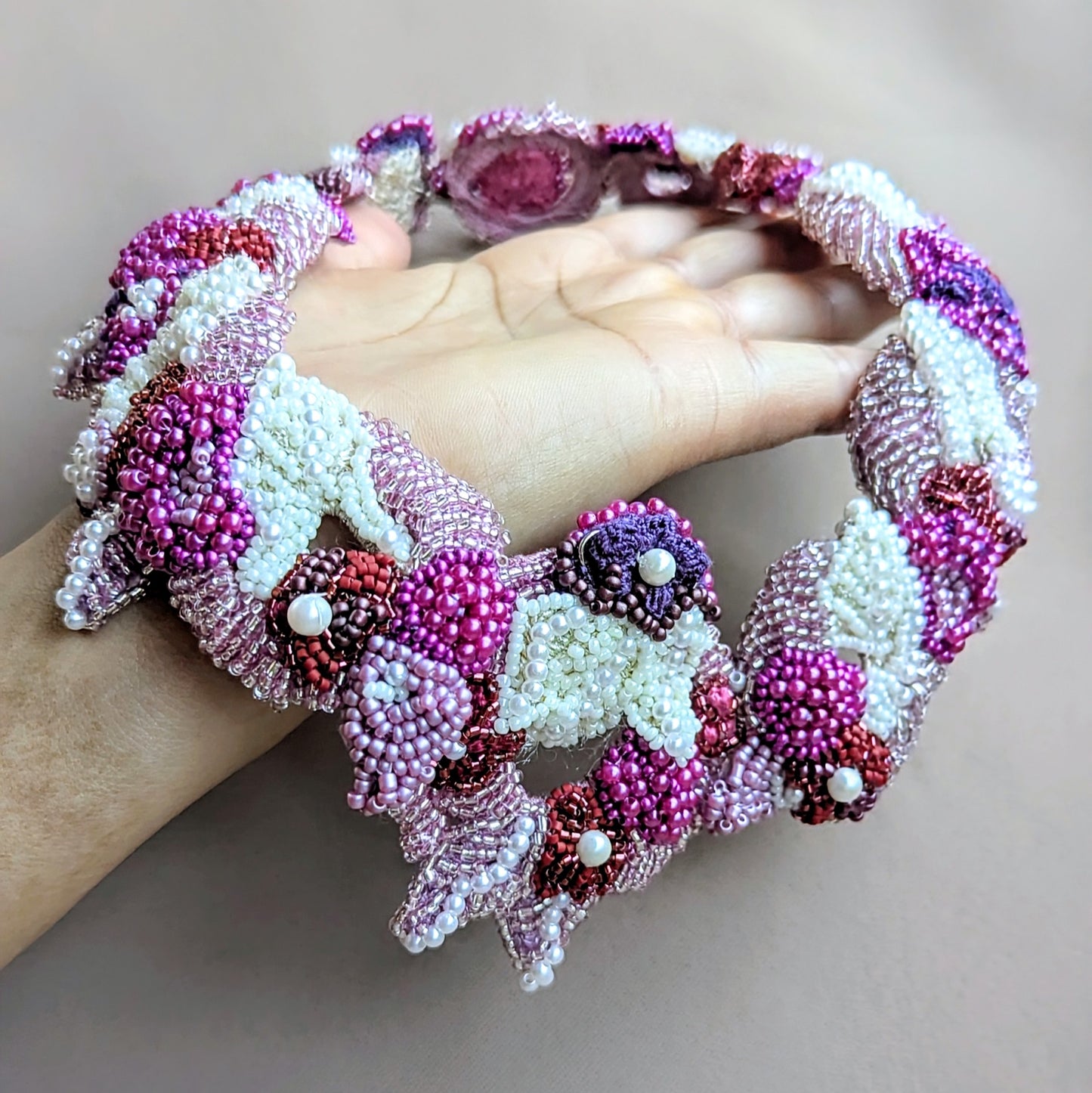 A handcrafted one-of-a-kind Indian bridal necklace featuring a detachable pendant crafted in elegant shades of purple, pink, white, and red displayed on an open palm against a neutral background. An ethically made fusion of bead embroidery, crochet and wirework designed for modern brides.