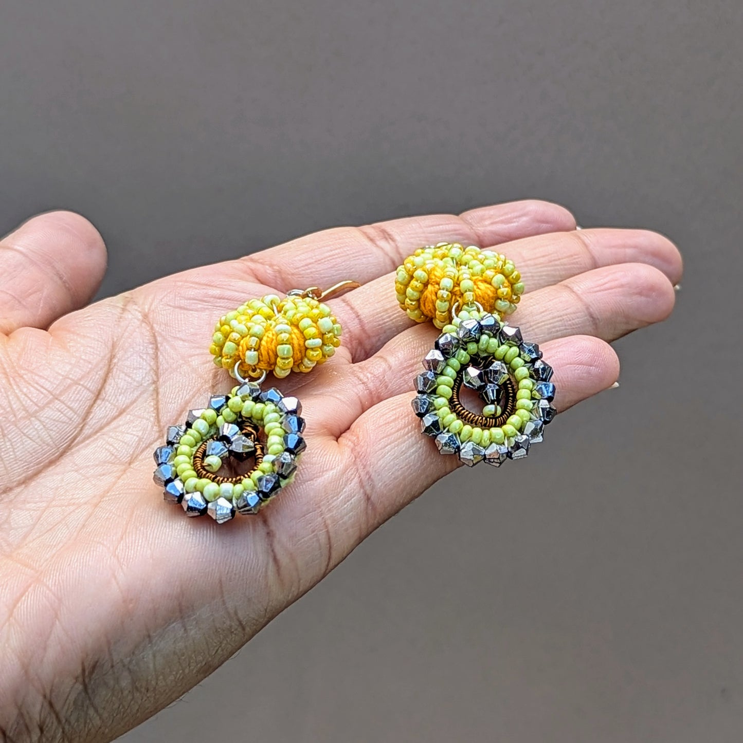 Hand holding a modern Indian-inspired fusion pair of artisan handcrafted earrings exclusive to Mayaani Jewellery UK. Featuring yellow and gunmetal grey tones of beadwork, hand crochet and wire wrapping.