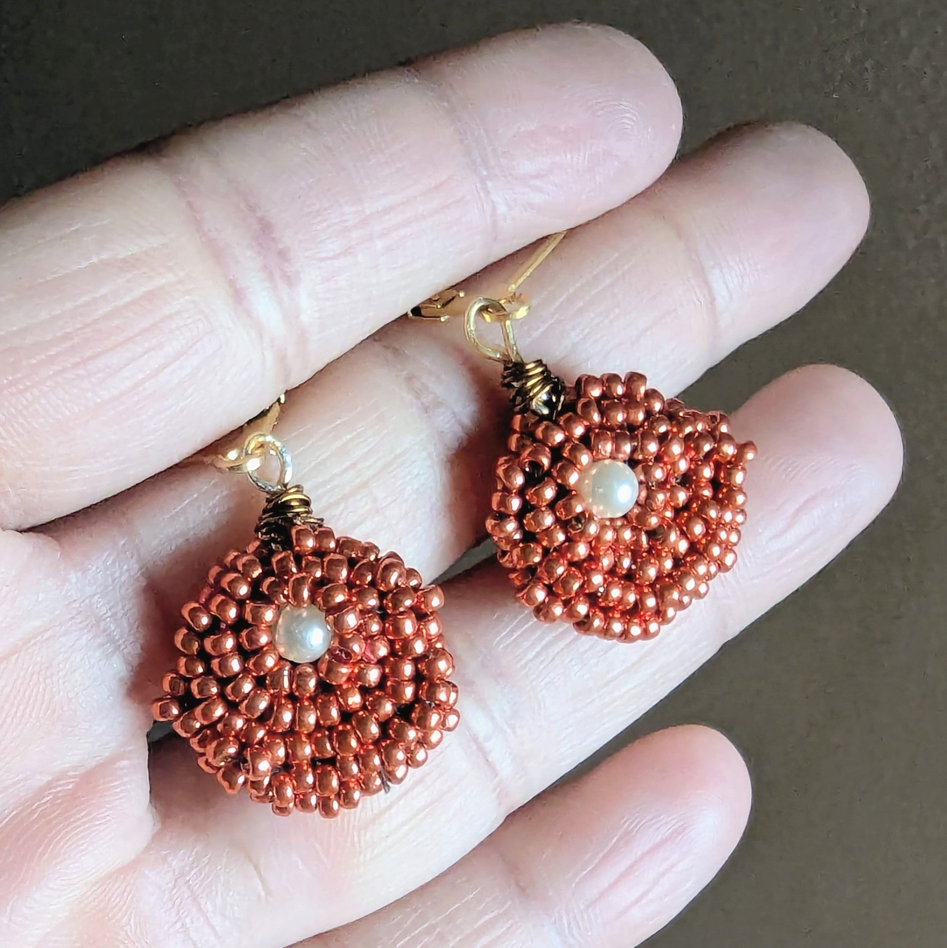 Hand holding a pair of artisan made copper and pearl seed bead earrings with gold stainless steel clasps designed in the UK.