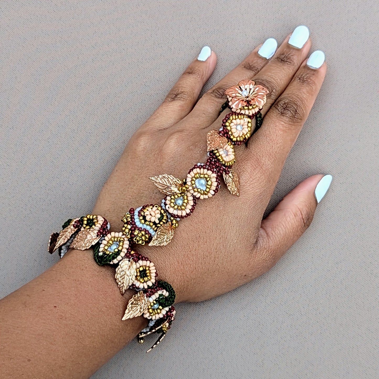 Hand wearing a decorative red, green, gold Indian hand bracelet featuring gold plated leaf details. This piece is a 3-in-1 early prototype of the signature Mayaani 5-in-1 Indian hand bracelet.