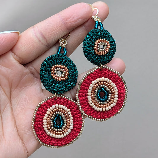 Hand holding a vibrant one-of-a-kind red, green & gold pair of handcrafted bead crochet earrings exclusive to sustainable brand Mayaani Jewellery UK. Inspired by auspicious Indian colours and the festival of Diwali, heritage is beautifully rewritten in this  statement making pair.