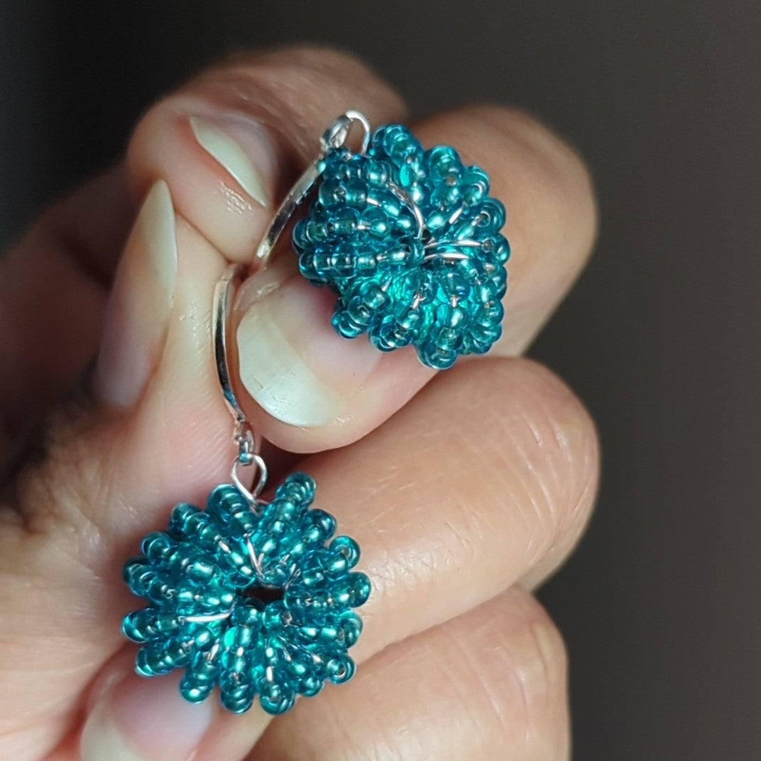 Close-up of artisan-made blue beaded earrings with sterling silver clasps and rose gold wire against a dark grey background.