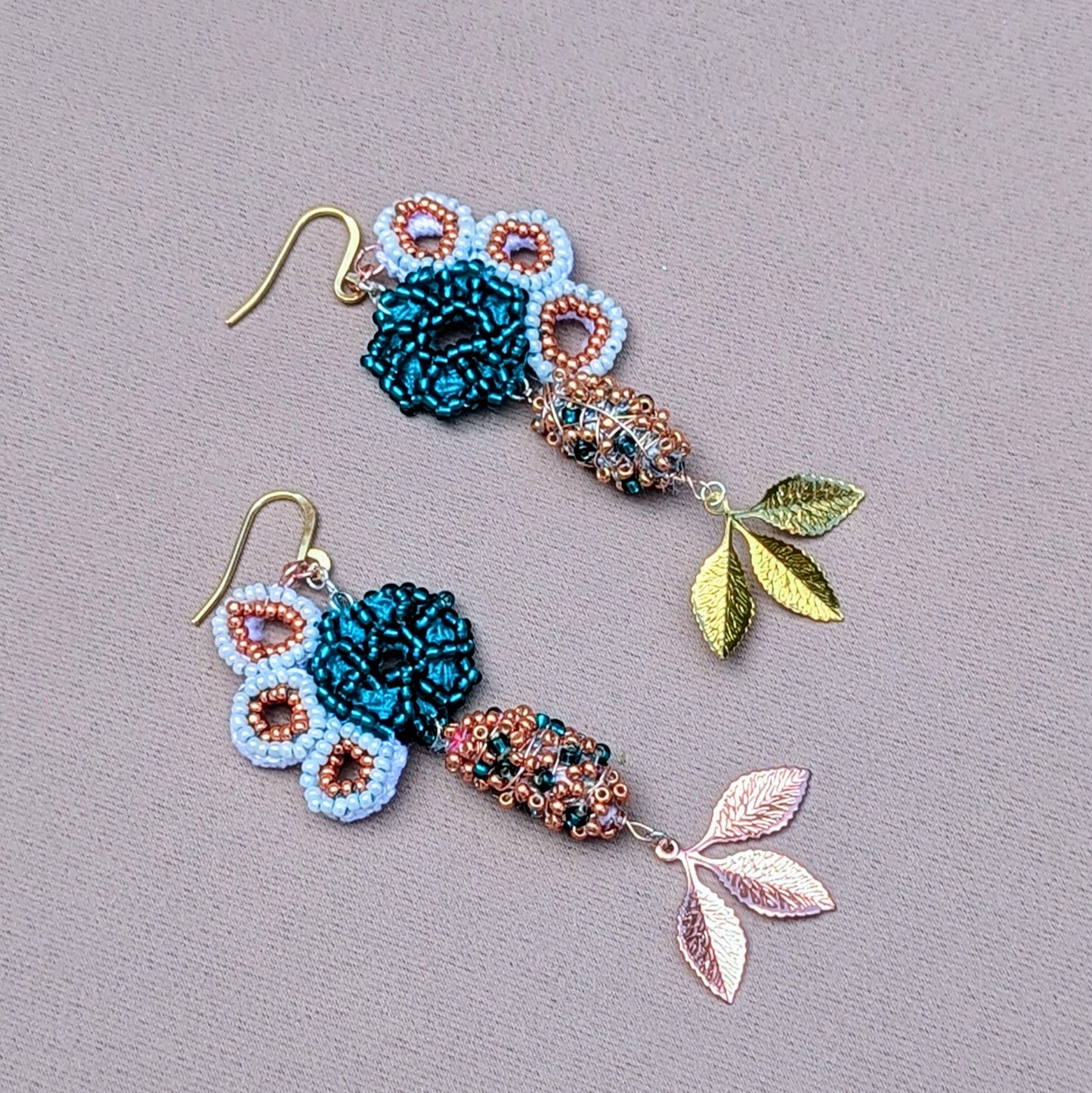 A handcrafted pair of floral abstract seahorse motif earrings on a grey background. Exclusive to Mayaani Jewellery UK this asymmetric pair features high quality white, emerald and rose gold seed beads, a signature design pair of scrap beads and two gold/ rose gold plated leaf charms.