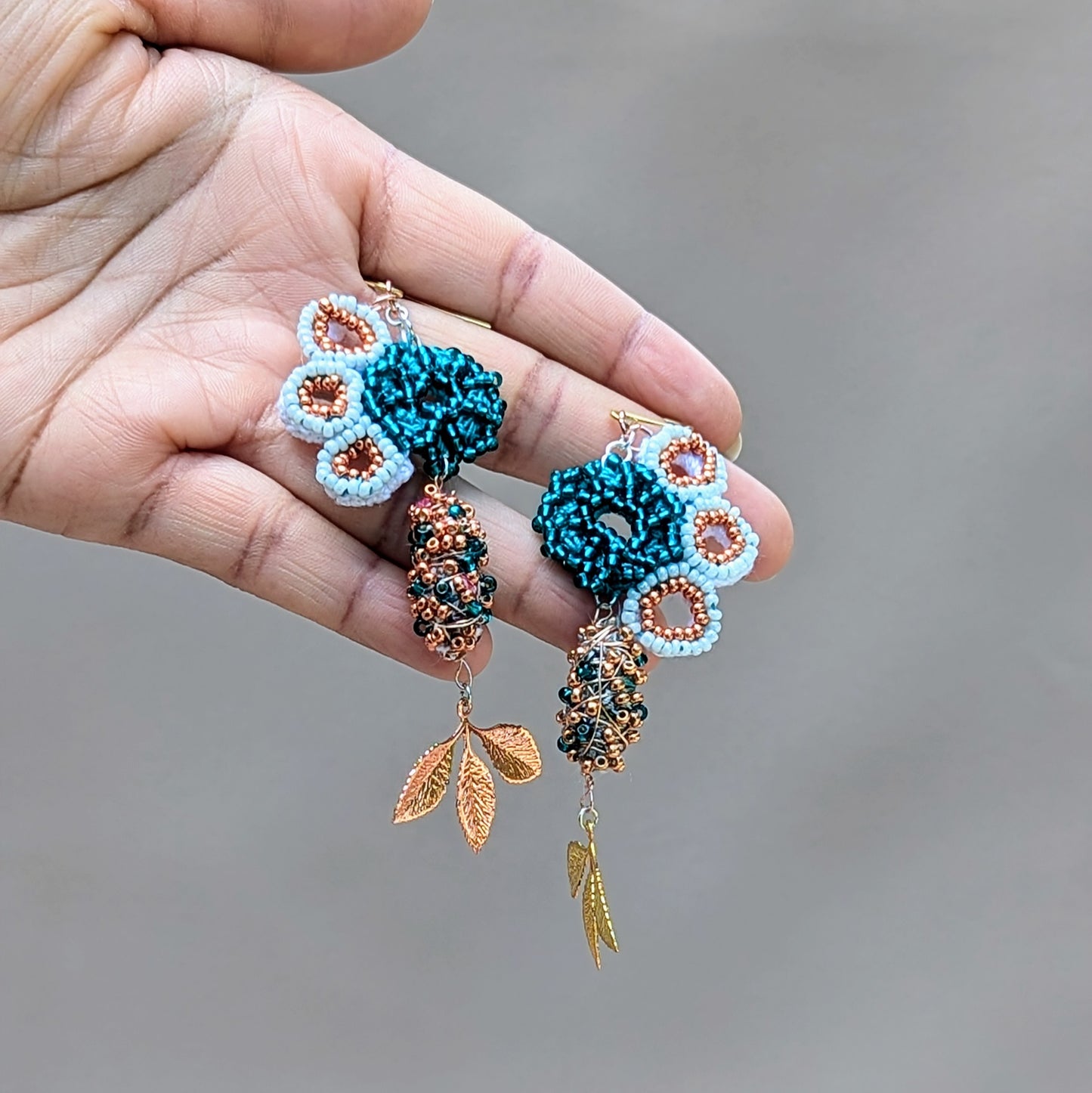 Hand holding an asymmetric scrap bead pair of Mayaani Jewellery earrings handcrafted in the UK using high quality glass seed beads in white, emerald green and rose gold alongside beads created using scraps from the jewellery making process.