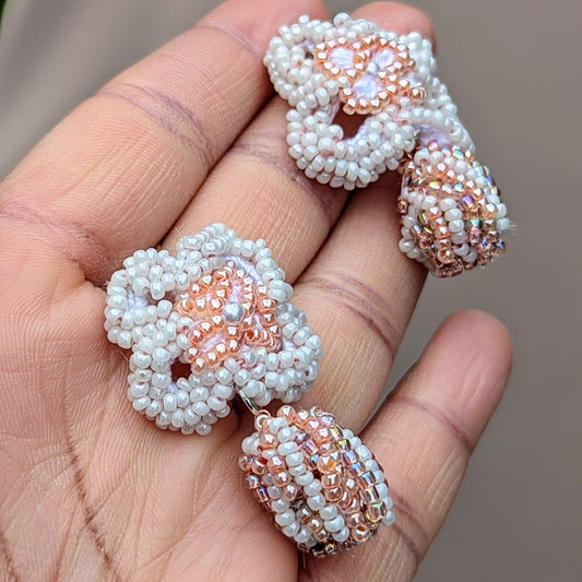 A hand holding an artisan handcrafted pair of peach and white bridal earrings by sustainable brand Mayaani Jewellery UK. Set against a soft neutral background, the earrings feature floral crochet motifs and a unique scrap bead drop.