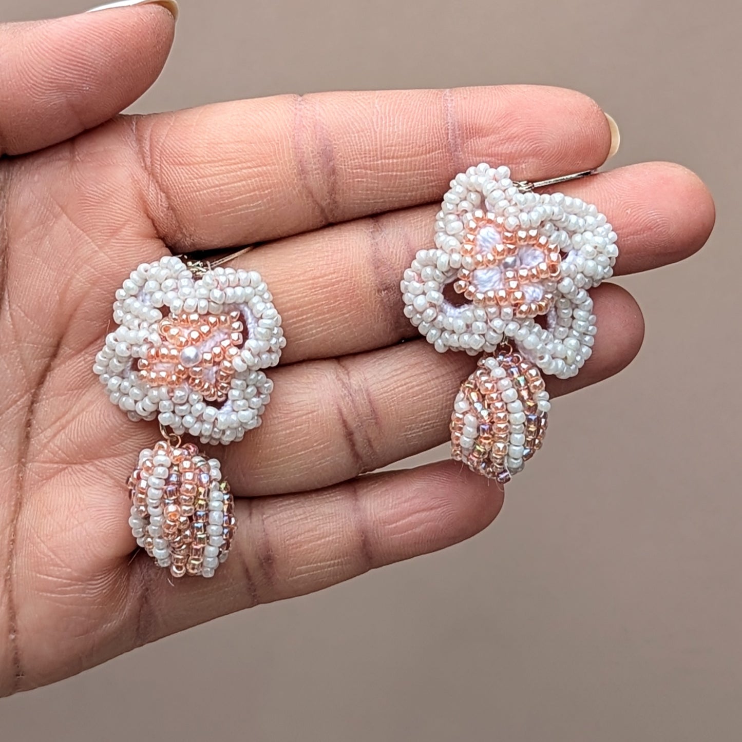 Flat lay of handcrafted white and peach floral earrings resting on a decorative white peacock trinket tray. Designed by Mayaani Jewellery UK, the pair features seed bead crochet, a central scrap bead, and sterling silver hooks - capturing eco-conscious bridal elegance.