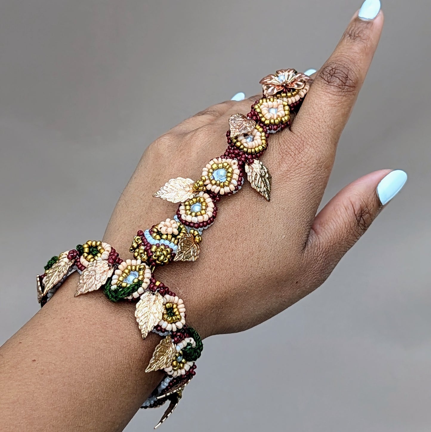 A woman's hand with white nail polish modelling an early prototype of the modern 5-in-1 red, green and gold Indian hand bracelet unique to Mayaani Jewellery featuring gold plated leaves.