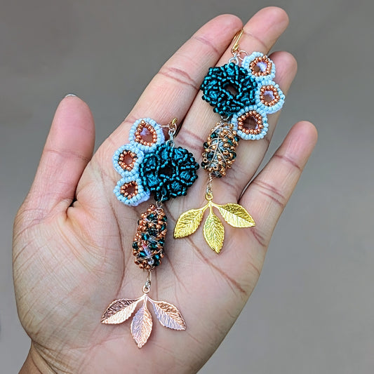 A hand holding a Mayaani Jewellery artisan handcrafted pair of decorative seahorse motif inspired earrings featuring emerald green, rose gold, and white seed beads coupled with rose gold and gold-plated leaves.
