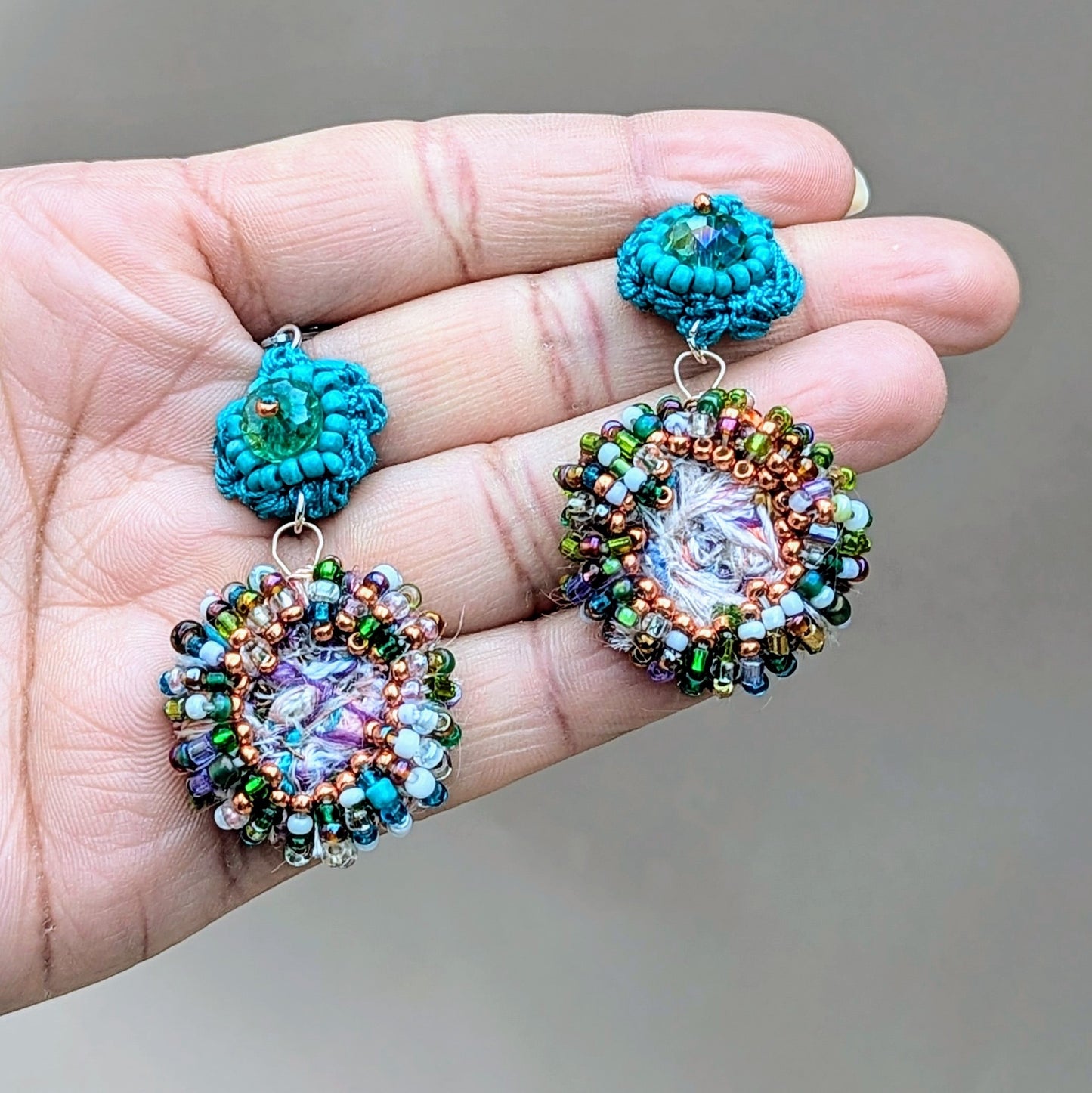 A hand displays a one-of-a-kind pair of statement earrings by Mayaani Jewellery UK, made from upcycled scrap beads in turquoise, green, purple, rose gold, and blue - showcasing a zero-waste, sustainable Indian design.