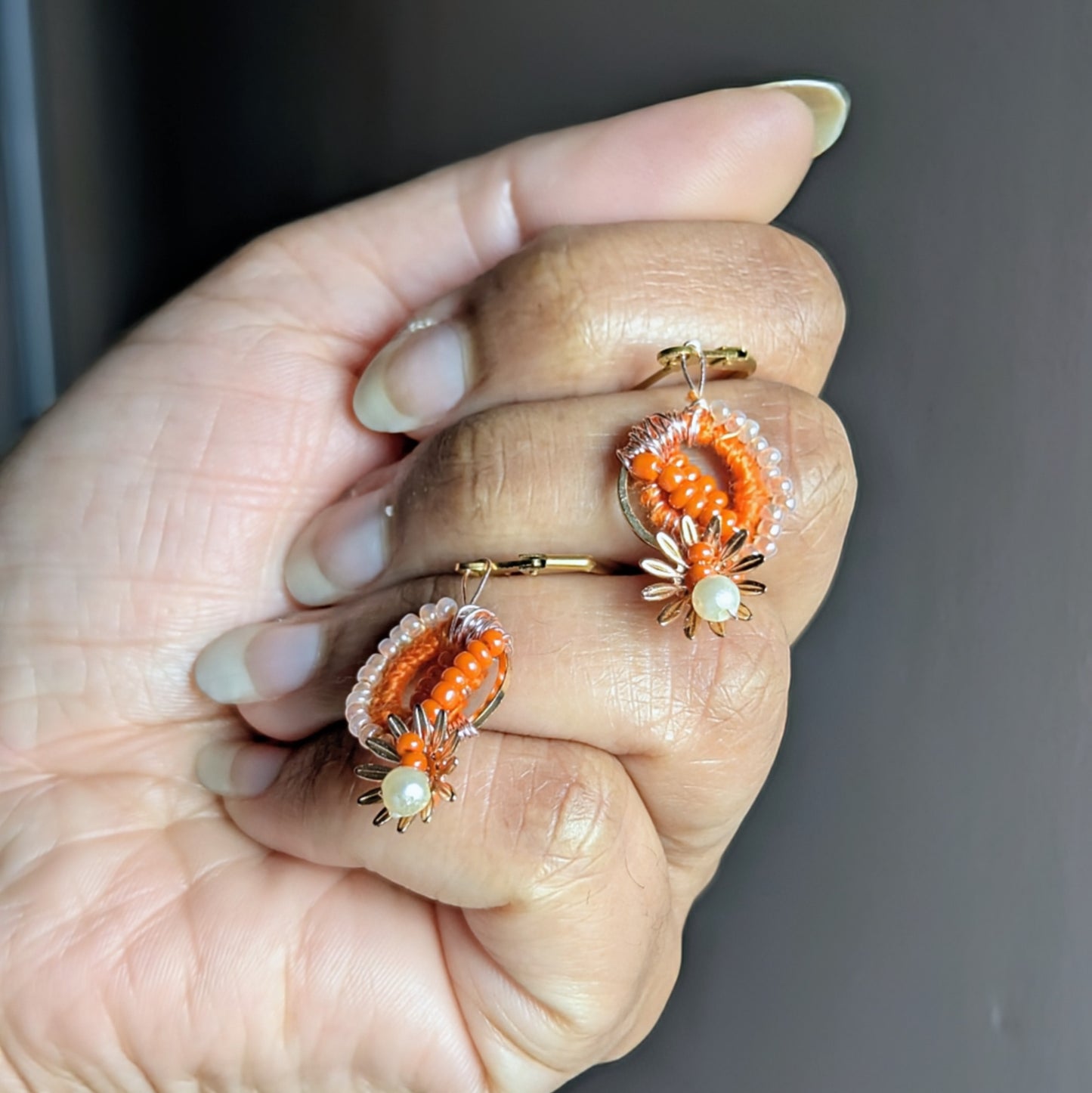 Hand holding artisan-made orange bead earrings with crochet and rose-gold wire details.