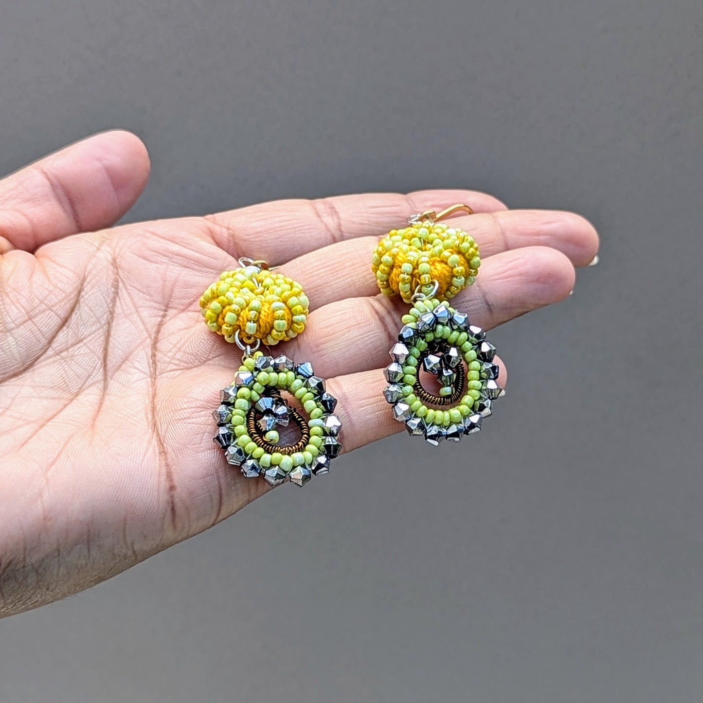 A pair of handcrafted yellow and gunmetal grey earrings held in the open palm of a hand against a neutral grey backdrop. Indian-inspired they are artisan handcrafted exclusively for Mayaani Jewellery UK.