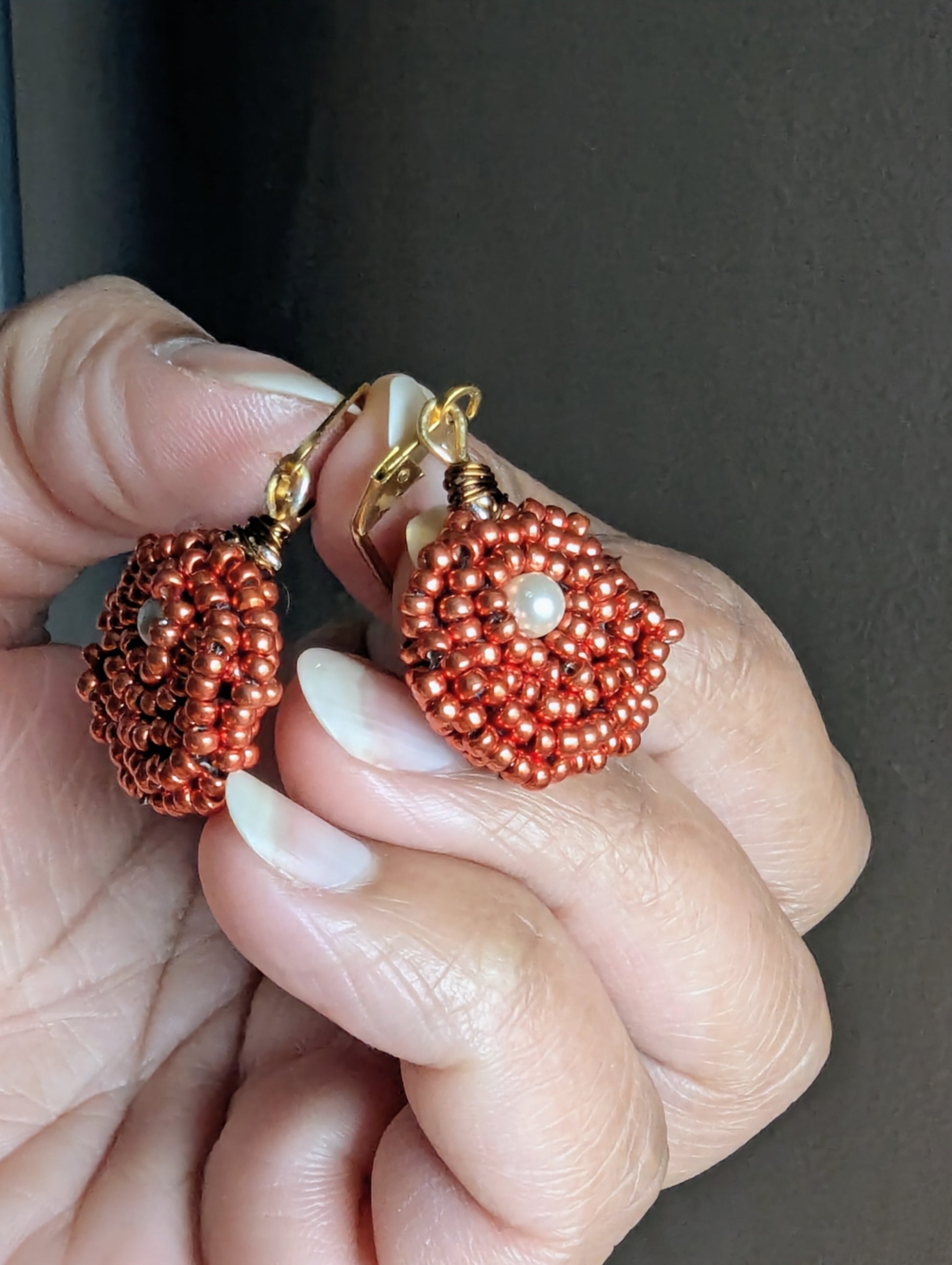 Hand holding a pair of gold clasp, copper and pearl seed bead earrings exclusively designed and made by Megha.