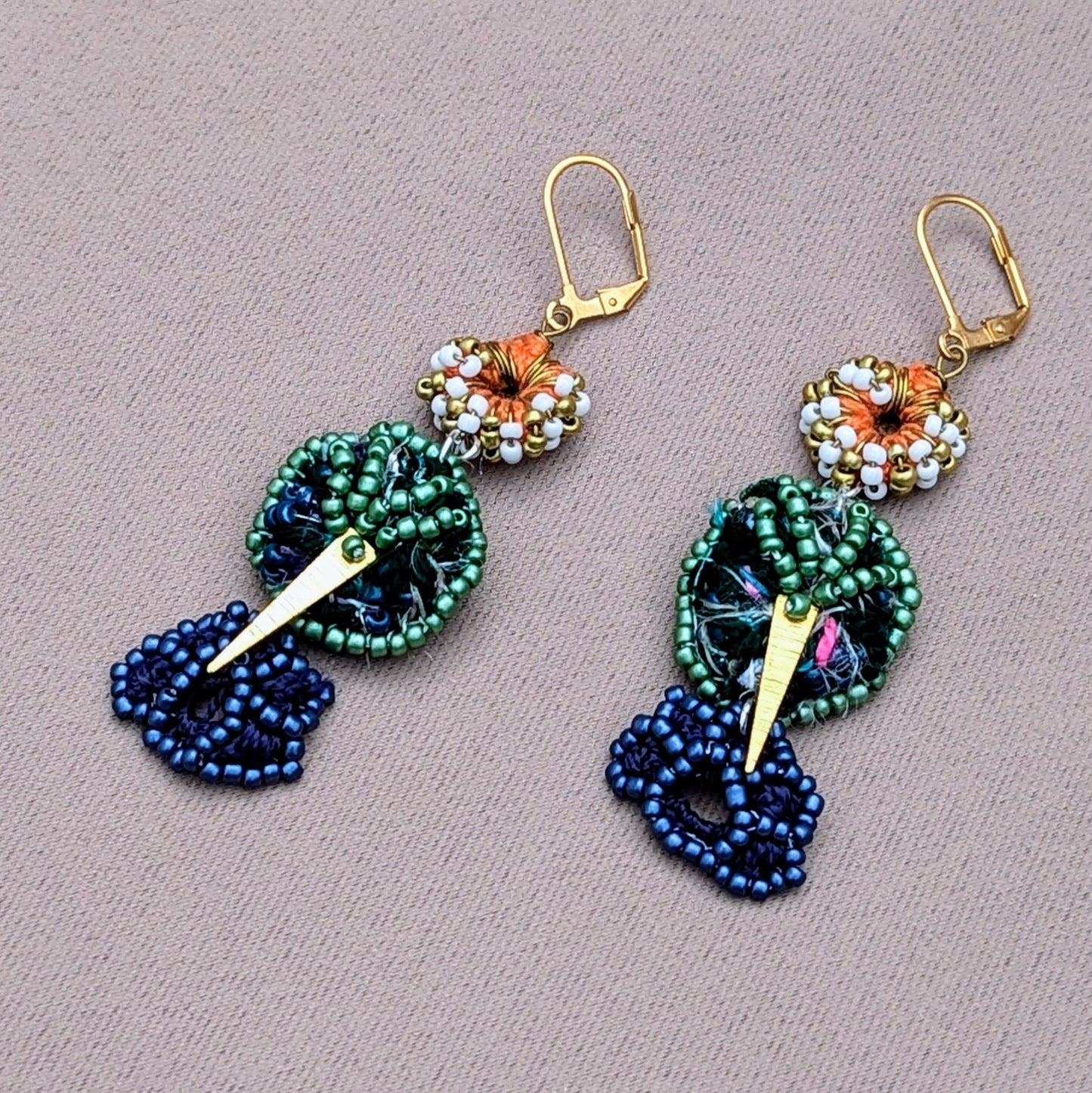 A sustainably handcrafted pair of artisan Indian statement earrings exclusive to Mayaani Jewellery UK. Featuring 18K gold triangle charms, hand crochet, bead and wire work in a mixture of greens, orange, blues, white and gold including an 18K gold triangle charm.