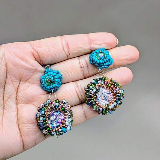 A hand holding a sustainably handcrafted pair of artisanal Indian inspired turquoise scrap bead earrings from Mayaani Jewellery UK, against a neutral grey backdrop. Featuring high quality upcycled seed beads in purple, green, blue, rose gold, and turquoise.