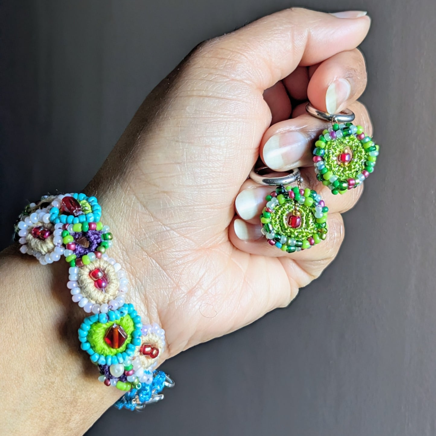 Hand holding a handcrafted pair of beaded earrings to compliment a modelled artisan made bracelet both highlighting crochet texture, mixed glass seed beads, and seed pearl accents.