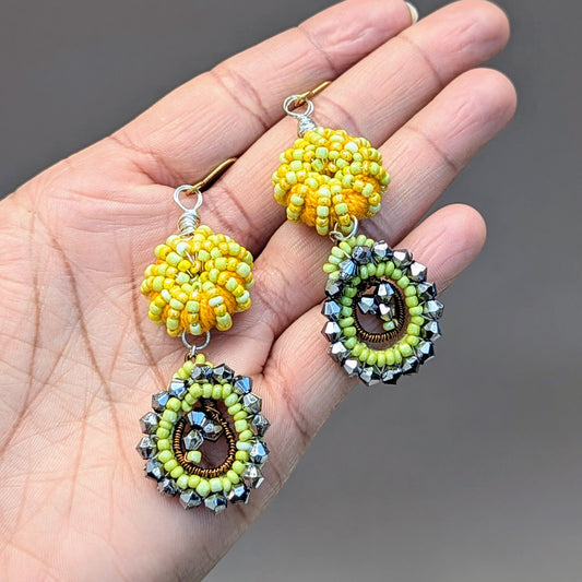 A pair of handmade yellow and gunmetal grey earrings featuring a circular crochet top with yellow seed beads and a teardrop pendant wrapped in antique copper wire. Exclusive to Mayaani Jewellery UK.