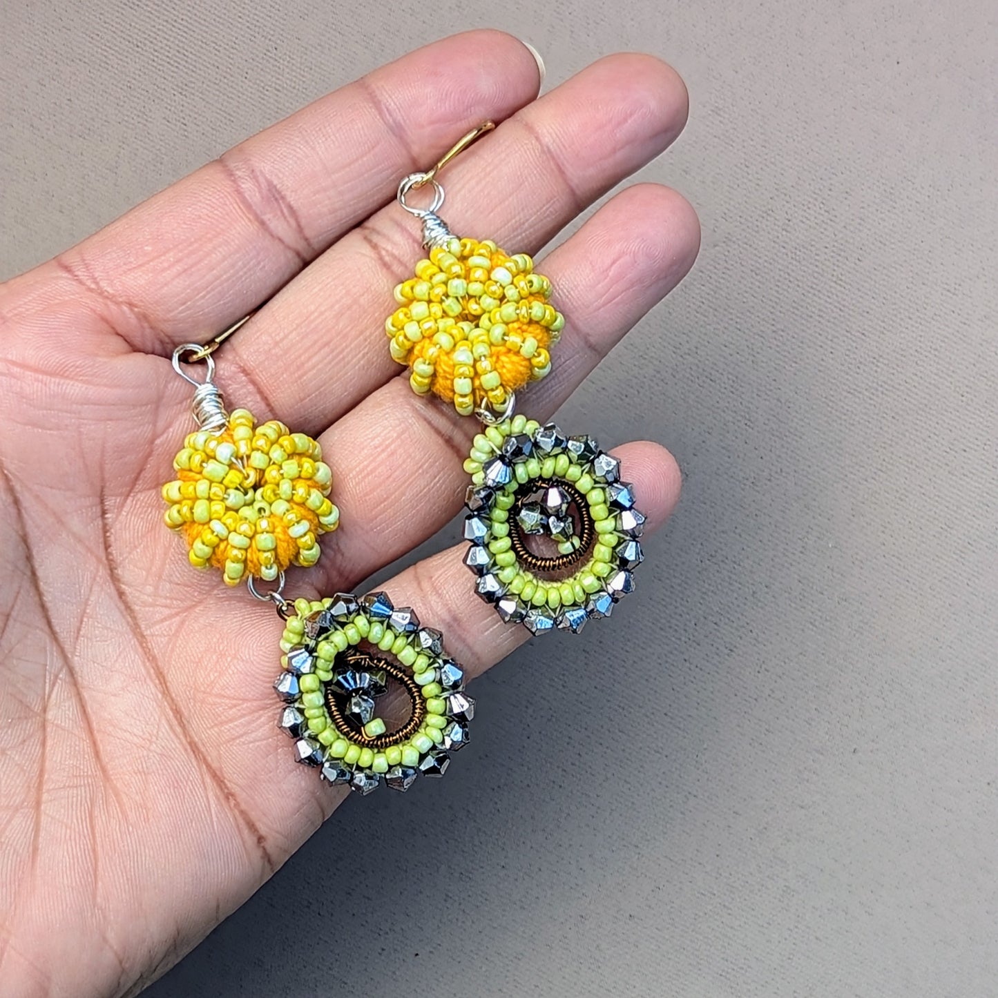 Hand holding an artisan handcrafted pair of woven bead, crochet and wire wrapped earrings in yellow and gunmetal grey exclusive to sustainable Indian brand Mayaani Jewellery UK.