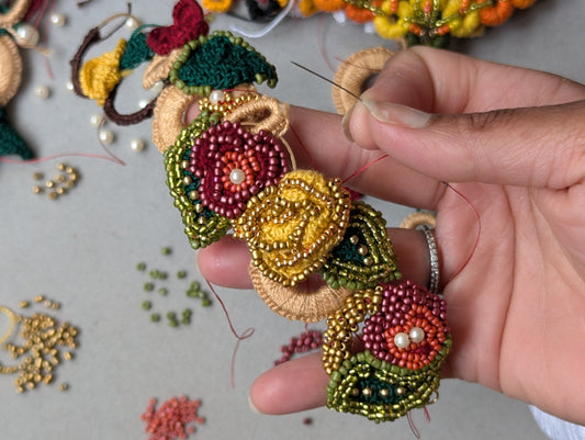 Flat lay of hands holding floral-inspired handmade jewellery in red, green, yellow and gold with pearl bead accents, on a neutral jewellery mat with tools in Mayaani Jewellery UK studio.