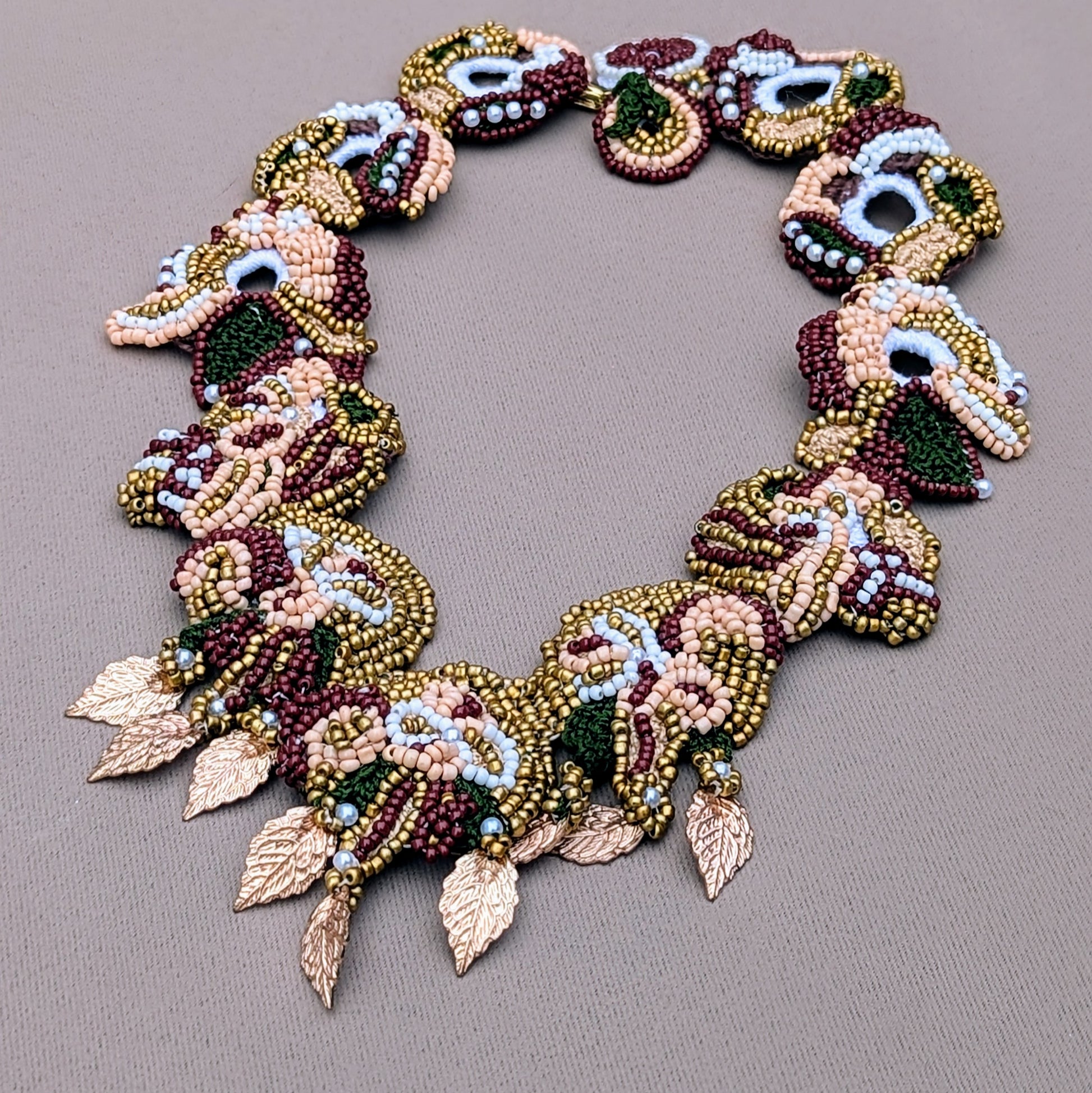 Flat lay of the Aishwarya Indian bridal couture necklace in red, green and gold handcrafted using Egyptian cotton crochet, premium seed beads and rose gold plated  leaves exclusively by Mayaani Jewellery UK. Pictured against a neutral grey backdrop.