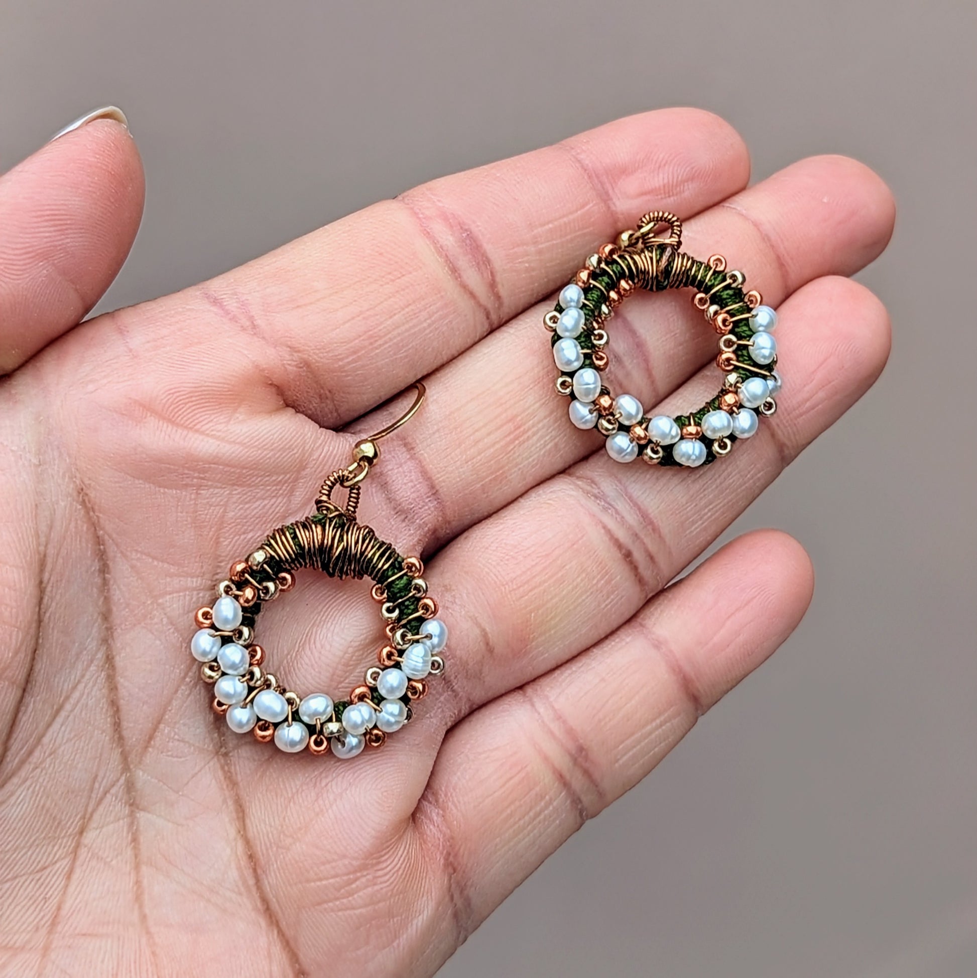 A unique limited edition design pictured in the palm of a hand. These artisan handcrafted earrings feature natural freshwater pearls and are exclusive to Mayaani Jewellery UK. Delicately crafted with green hand crochet, antique bronze wire wrapping, and gold beadwork arranged in a circular pattern, they are finished with gold plated stainless steel  earrings hooks. This Indian-inspired twist on classic pearl earrings makes them perfect for special occasions and beyond.