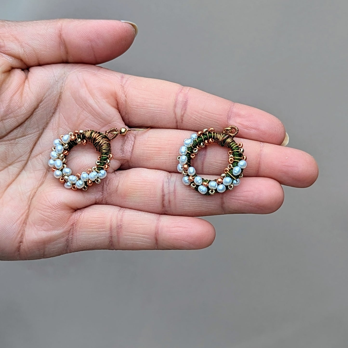 Mayaani Jewellery UK presents a limited edition, artisan-crafted design featuring natural freshwater pearls in the palm of a hand. These elegant earrings are adorned with delicate green hand crochet, antique bronze wire wrapping, and gold beadwork in a circular arrangement. Finished with gold-plated stainless steel hooks, they add an Indian-inspired touch to classic pearl jewellery.