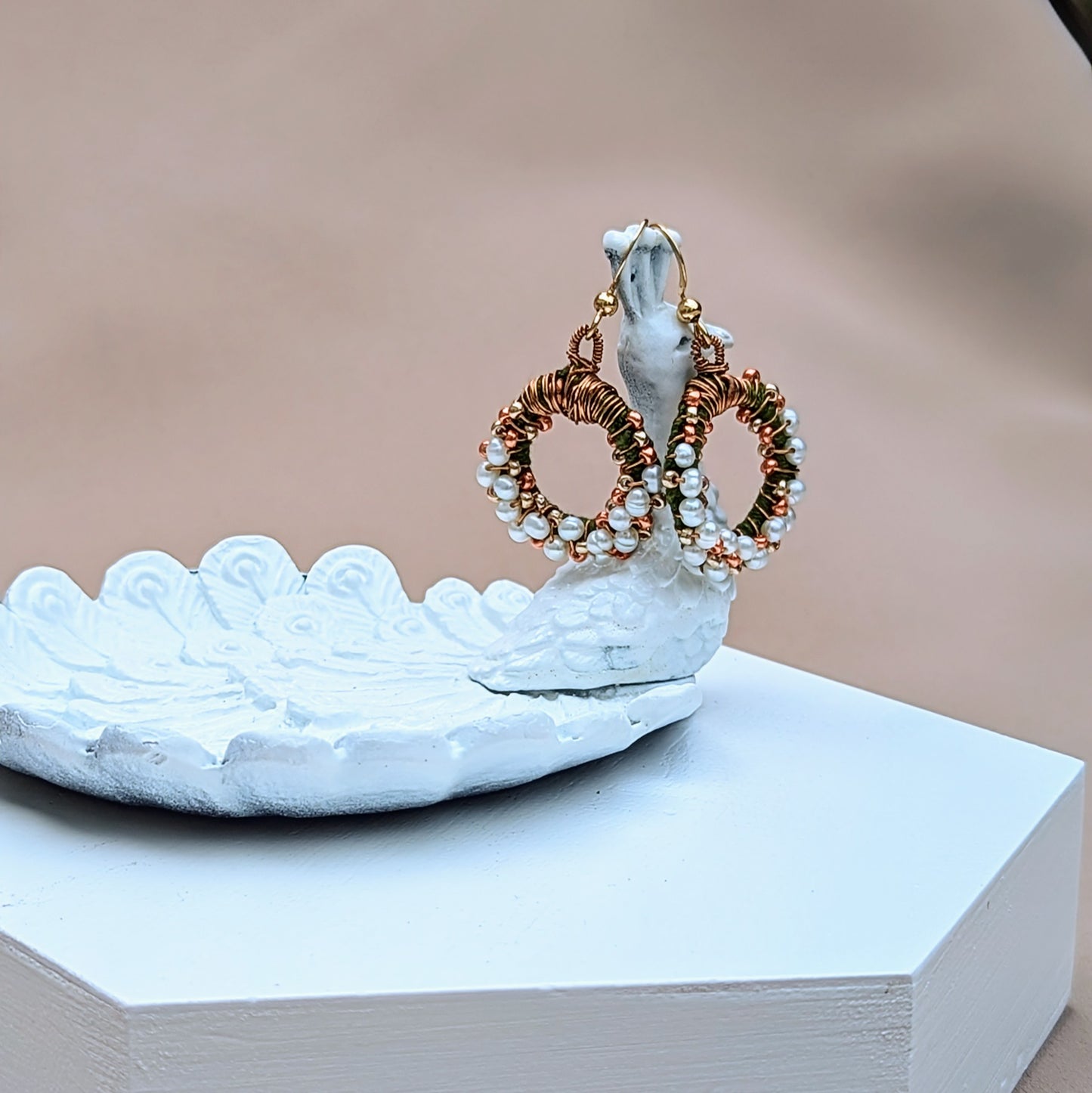 Displayed on a white peacock trinket dish, this limited edition design, exclusive to Mayaani Jewellery, features artisan-crafted natural freshwater pearl earrings with intricate green crochet, antique bronze wire wrapping and gold beadwork in a circular design. Finished with gold-plated stainless steel hooks, this Indian-inspired twist on pearl earrings is perfect for special events and more.