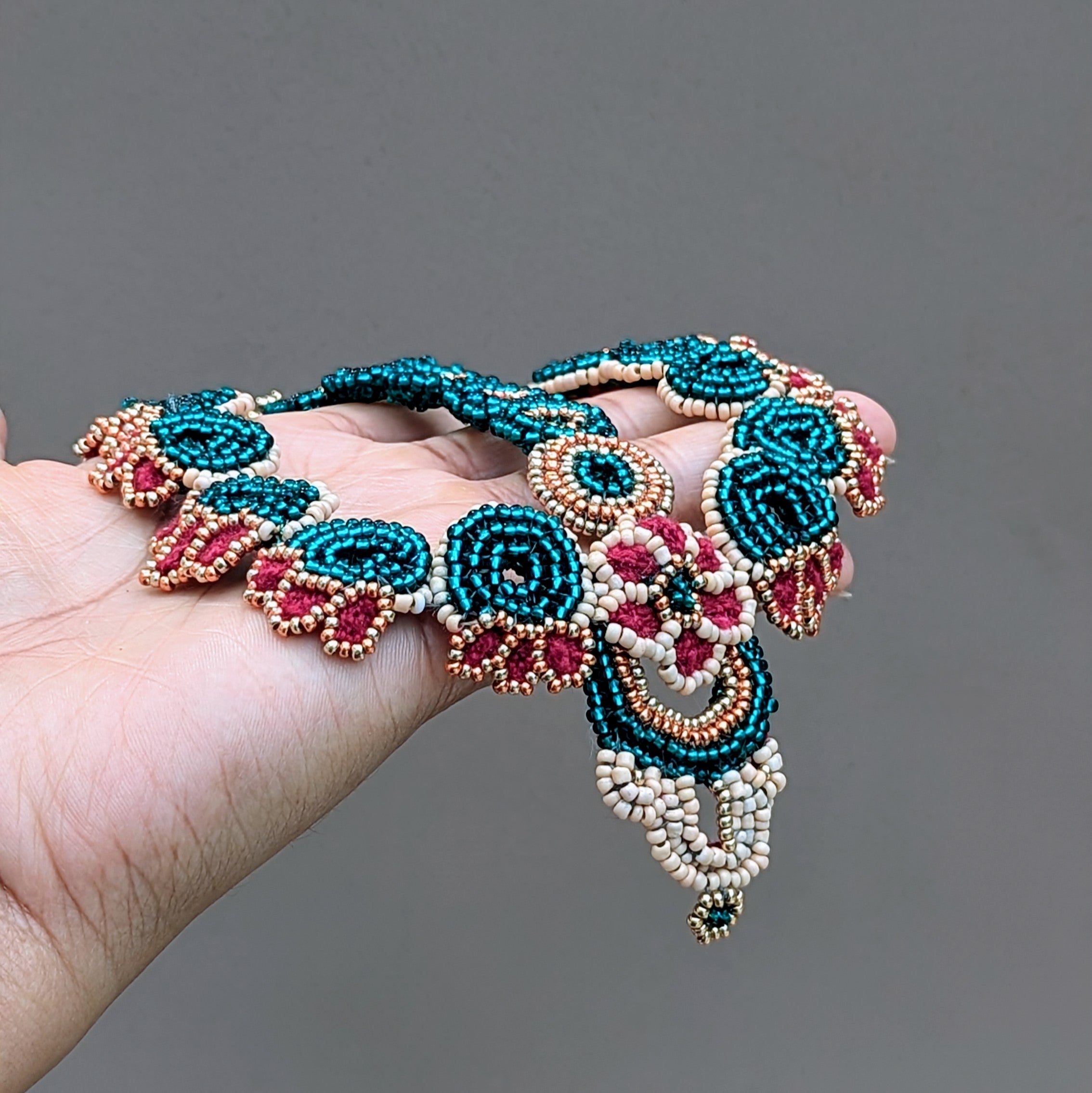 Hand holding a one of a kind Indian bridal headpiece exclusive to Mayaani Jewellery UK featuring delicate hand crochet elements and intricate hand bead work. The richness of red meets with the bold brilliance of emerald green accented with rose gold, gold and peach tones for a statement piece full of traditional artistry with an aesthetic perfect for the  modern Indian bride.