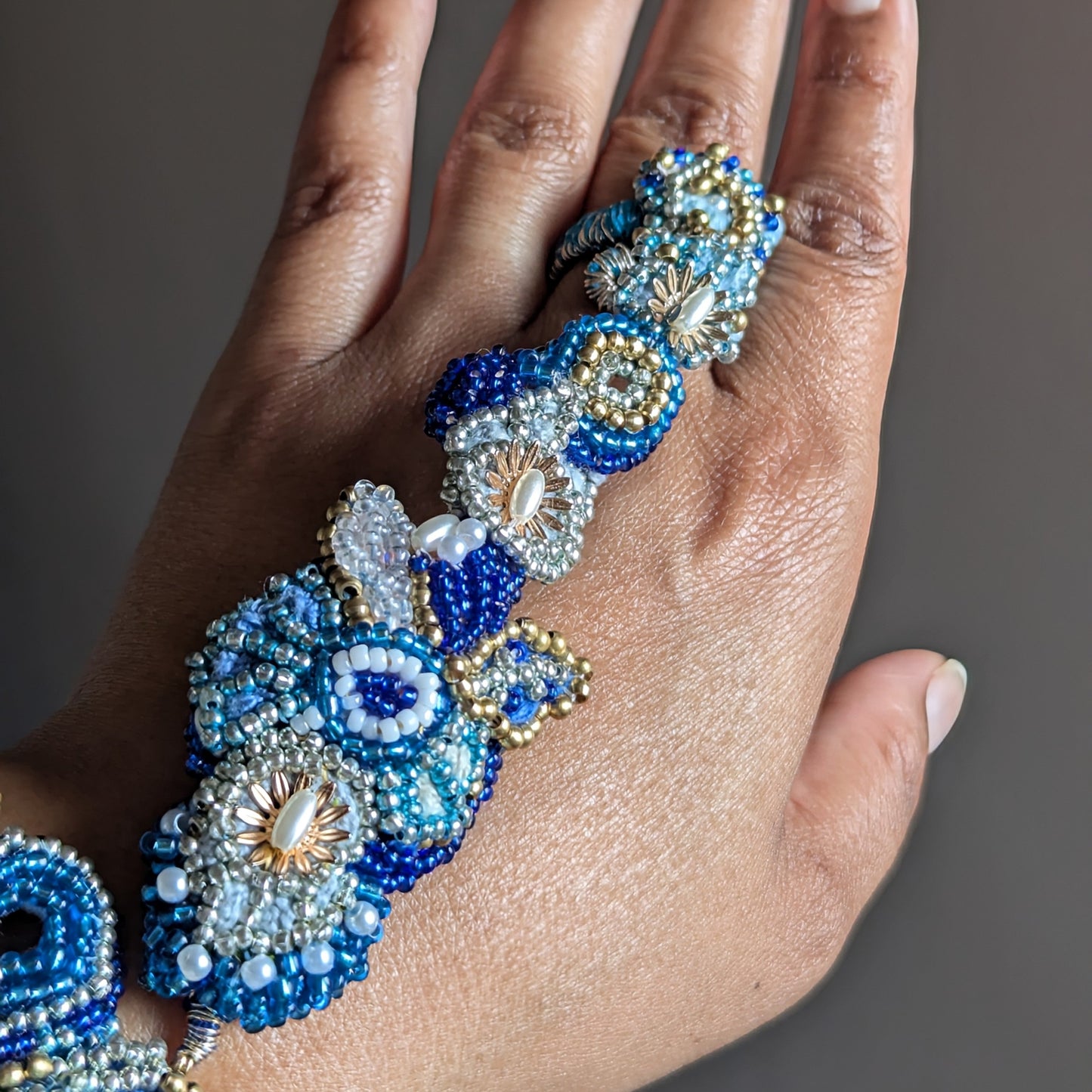 A close-up of a hand wearing the intricate Nira 5-in-1 beaded floral Indian hand bracelet featuring blue, white, and gold seed beads in a detailed floral patterns, set against a solid dark grey background.