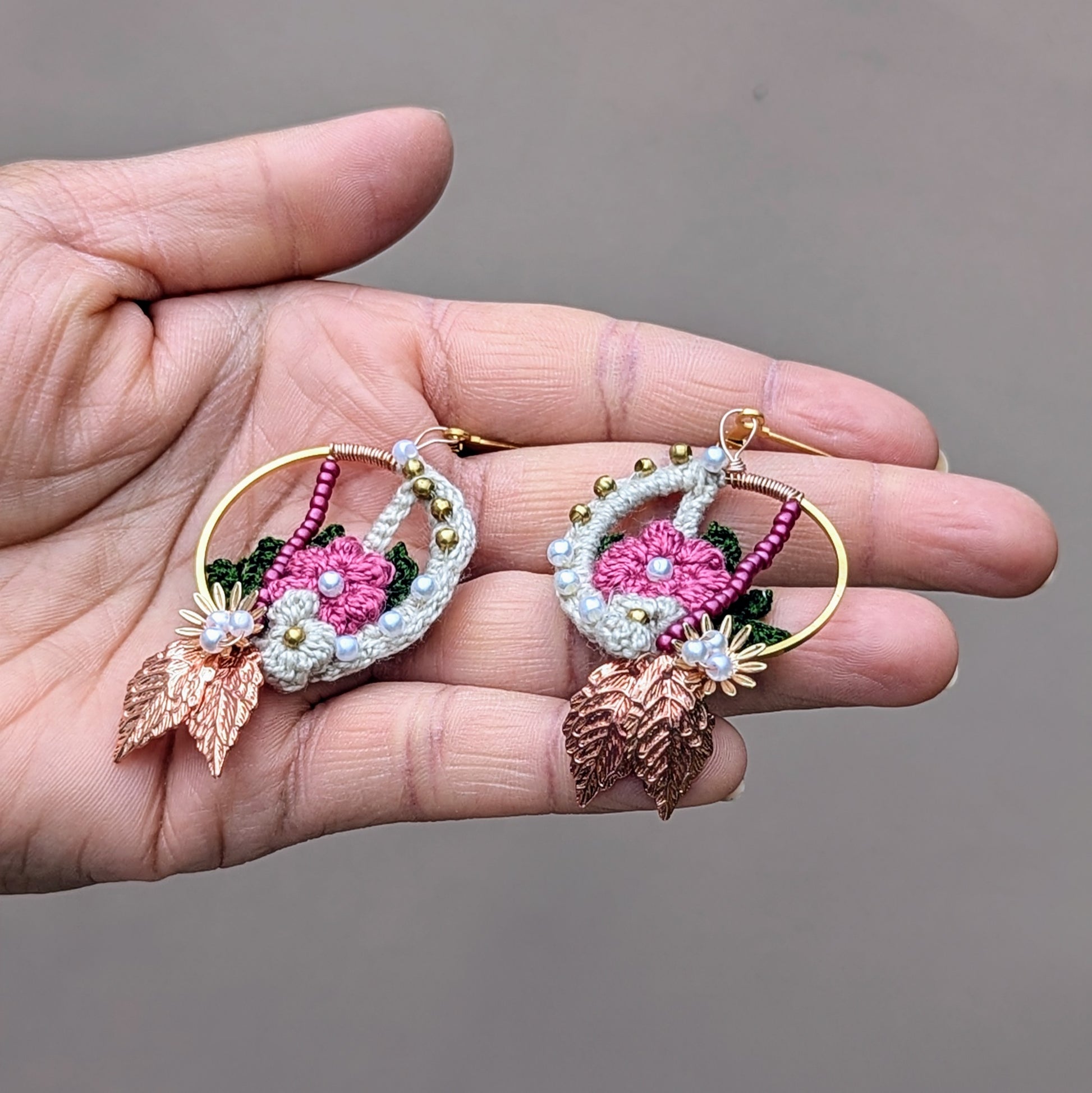 A pair of handcrafted Indian-inspired earrings pictured in a hand, from Mayaani Jewellery, a sustainable UK brand. These exclusive earrings showcase a beautiful mix of cream, pink, and dark green floral and leaf motifs, adorned with solid brass, pink, and seed pearl beads. Finished with rose gold leaves and gold-plated stainless steel clasps, they are lightweight and make a stunning statement. Perfect for weddings, special occasions, or everyday wear, they blend artistry and versatility seamlessly.