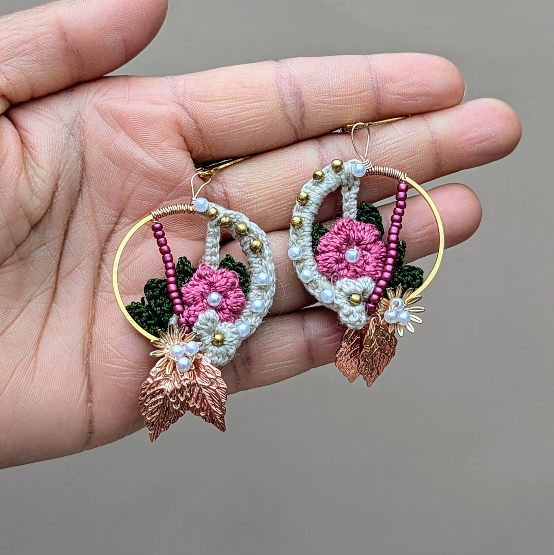Adorn yourself with handcrafted Indian-inspired earrings, held in the palm of a hand, from Mayaani Jewellery, a sustainable UK brand. Showcasing a blend of cream, pink, and dark green floral and leaf motifs, these earrings are embellished with solid brass, pink, and seed pearl beads. The design is completed with rose gold leaves and gold-plated stainless steel clasps, offering a lightweight and captivating accessory for weddings, special events, or everyday wear.